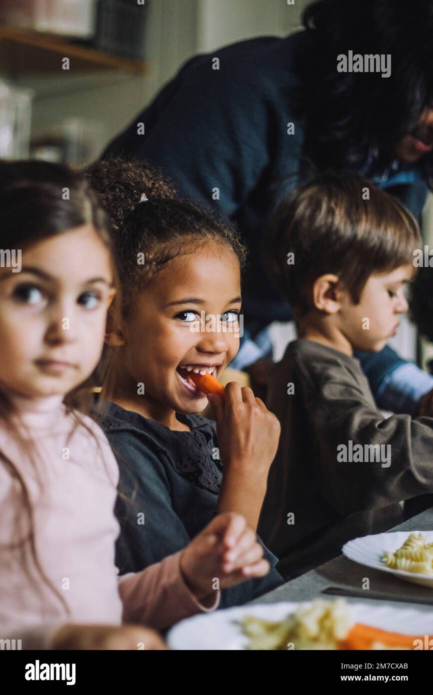 Happy girl eating carrot for breakfast with classmates in kindergarten