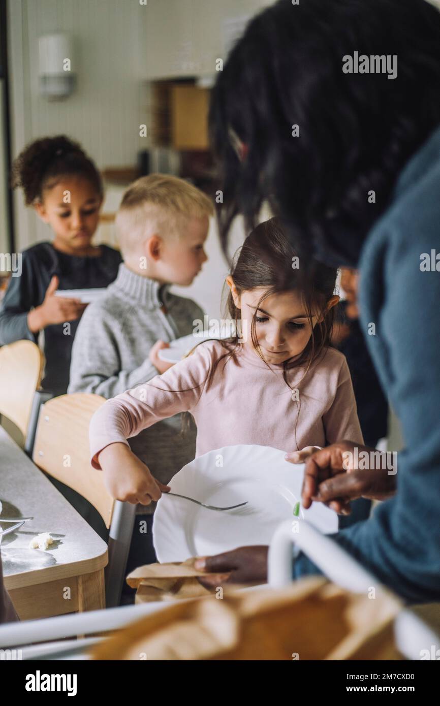 Girl throwing leftover food in paper bag held by female teacher at kindergarten Stock Photo