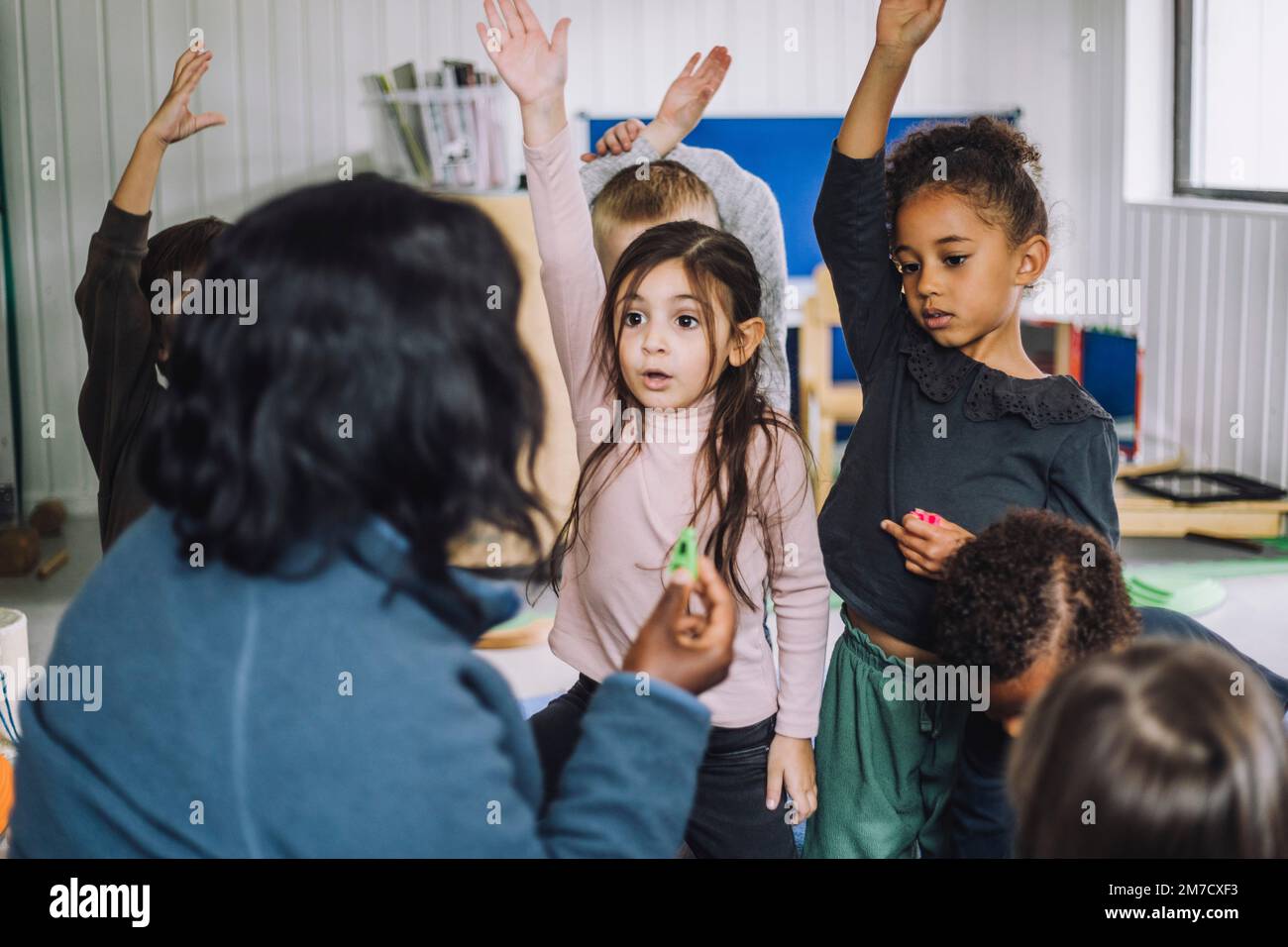 Female teacher asking questions and students raising hand to answer at day care center Stock Photo