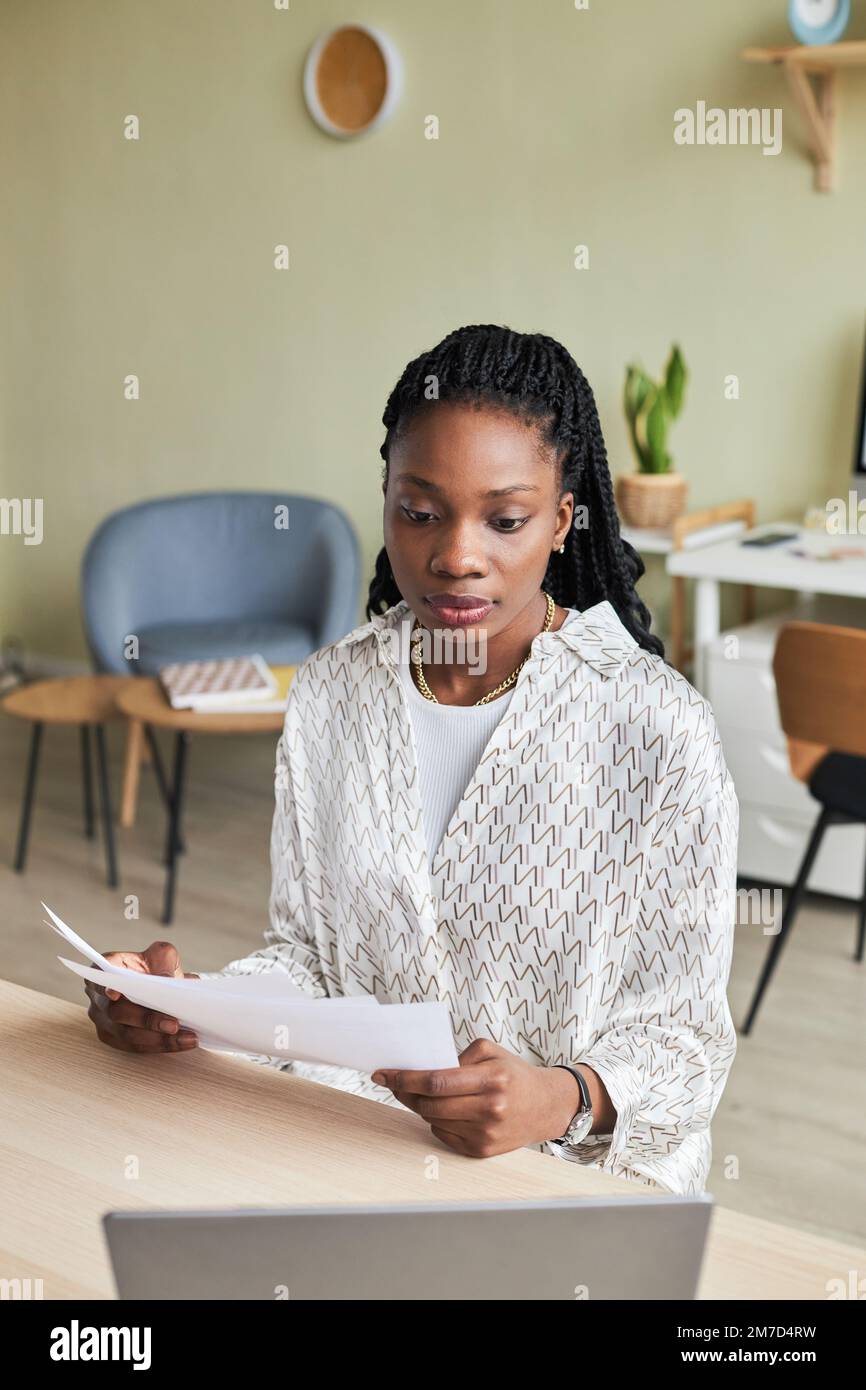 Vertical portrait of black young woman using laptop while working at desk in minimal office setting Stock Photo