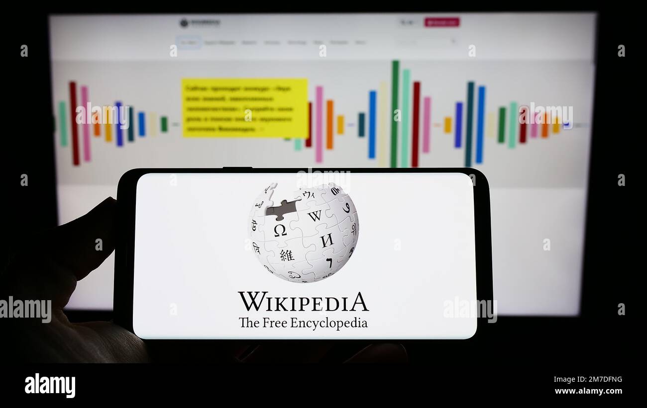 Person holding cellphone with logo of online encyclopedia Wikipedia on screen in front of Wikimedia website. Focus on phone display. Stock Photo