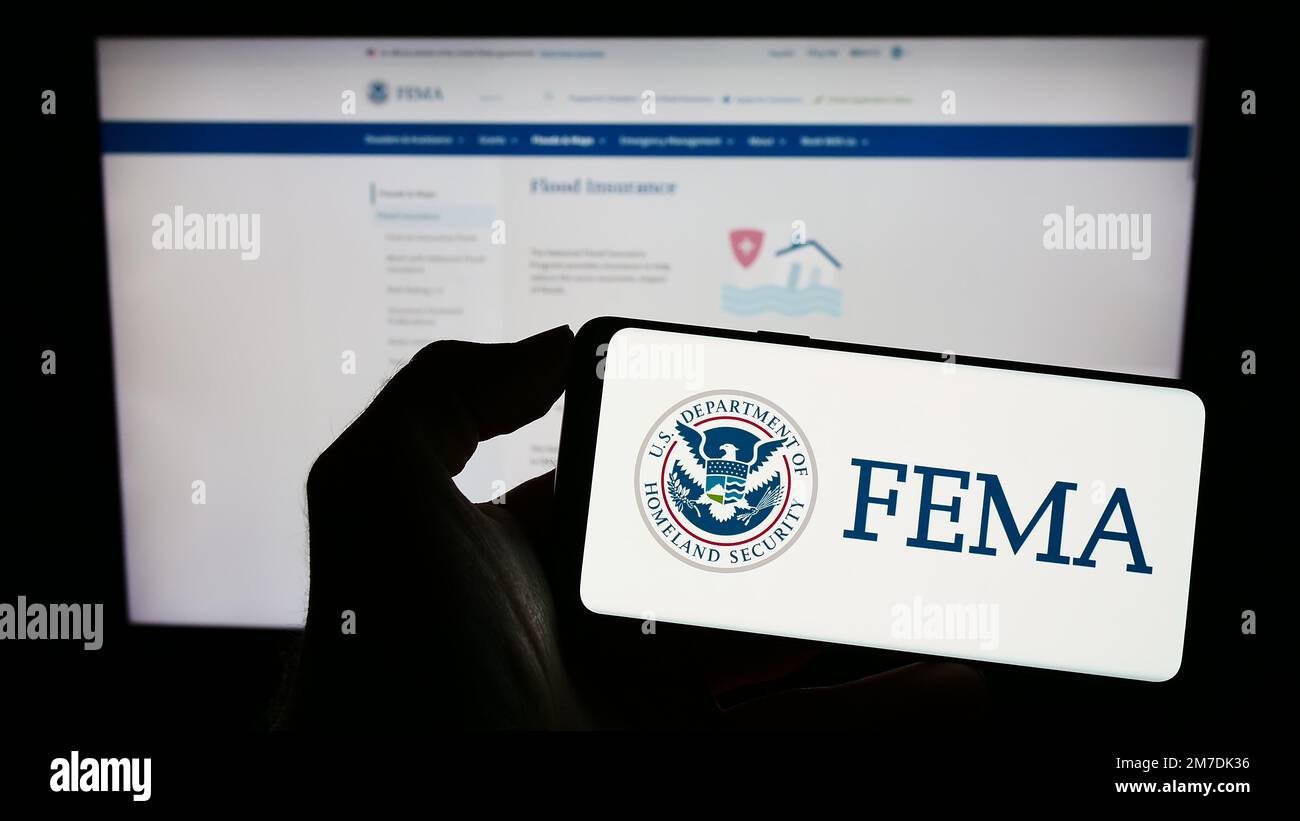 Person holding cellphone with seal of Federal Emergency Management Agency (FEMA) on screen in front of webpage. Focus on phone display. Stock Photo