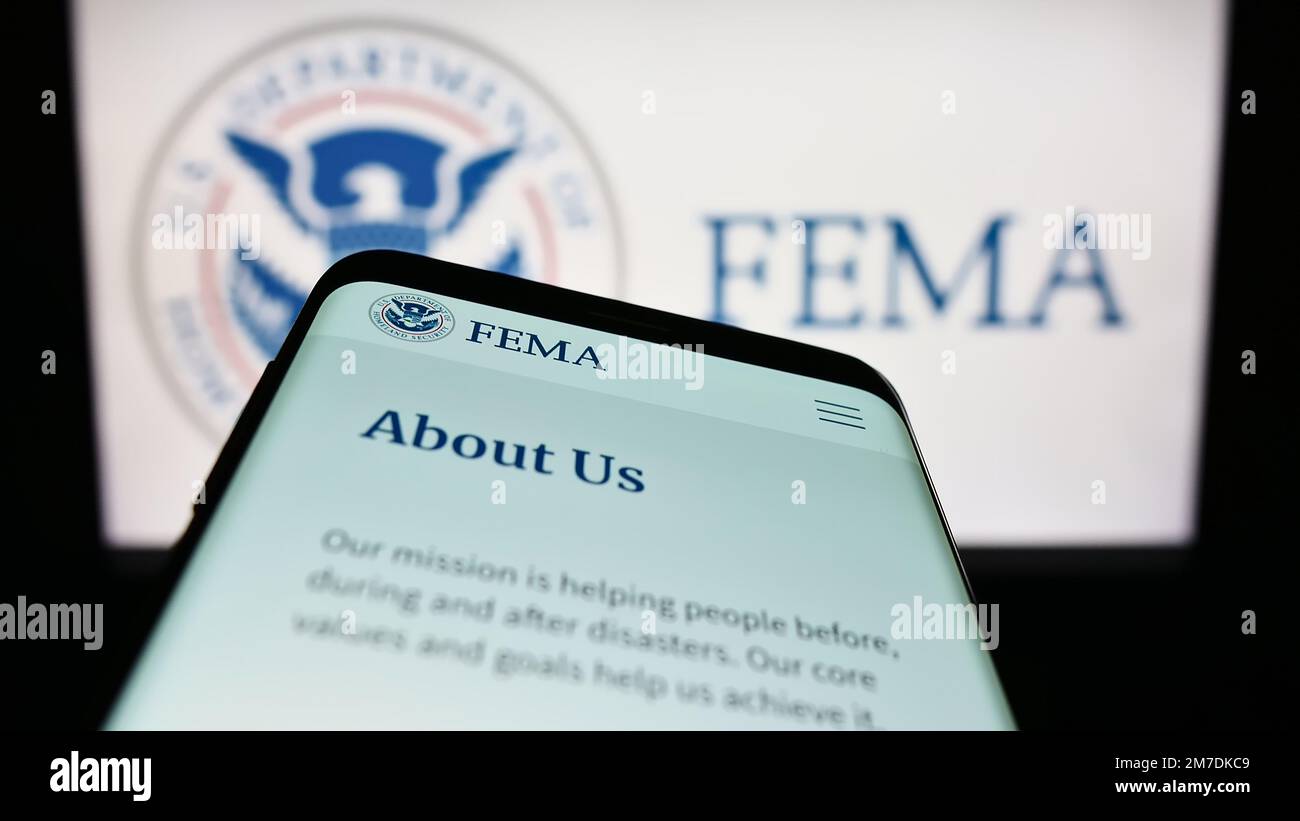Smartphone with website of Federal Emergency Management Agency (FEMA) on screen in front of seal. Focus on top-left of phone display. Stock Photo