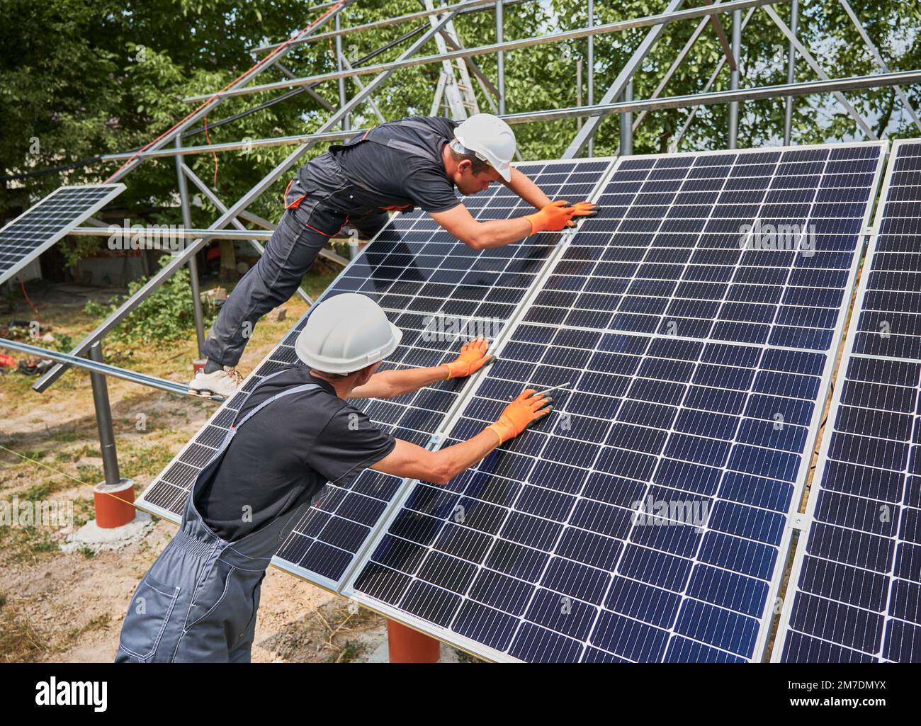 Workers installing photovoltaic solar panel system outdoors. Men engineers placing solar module on metal rails, wearing construction helmets and work gloves. Renewable and ecological energy. Stock Photo