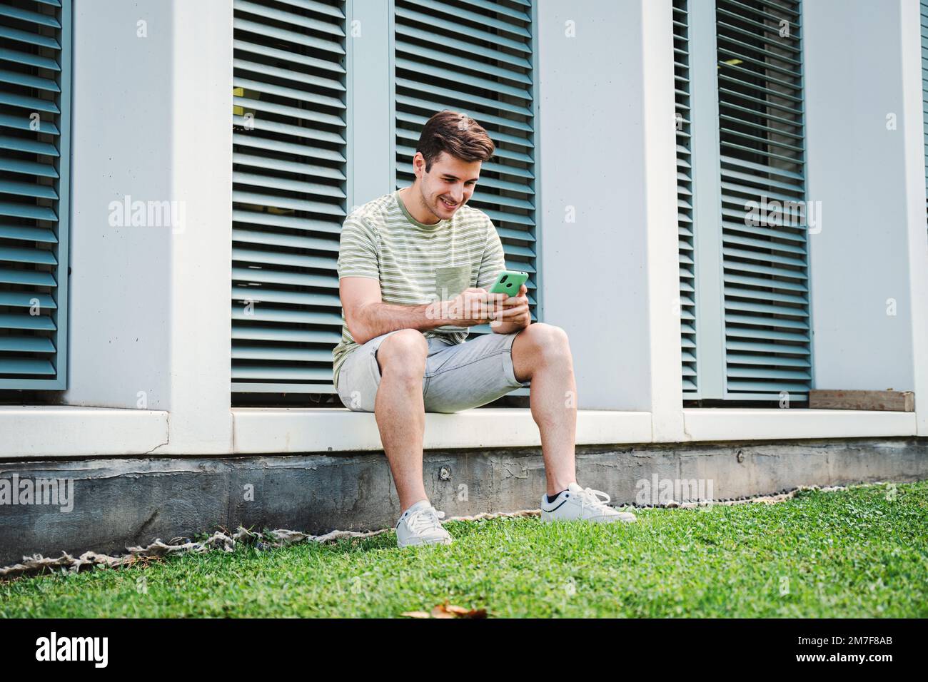 Young guy having fun playing a game with a cellphone sitting on the university campus after class. Teenage student smiling typing on a social media app with a smartphone device. High quality photo Stock Photo