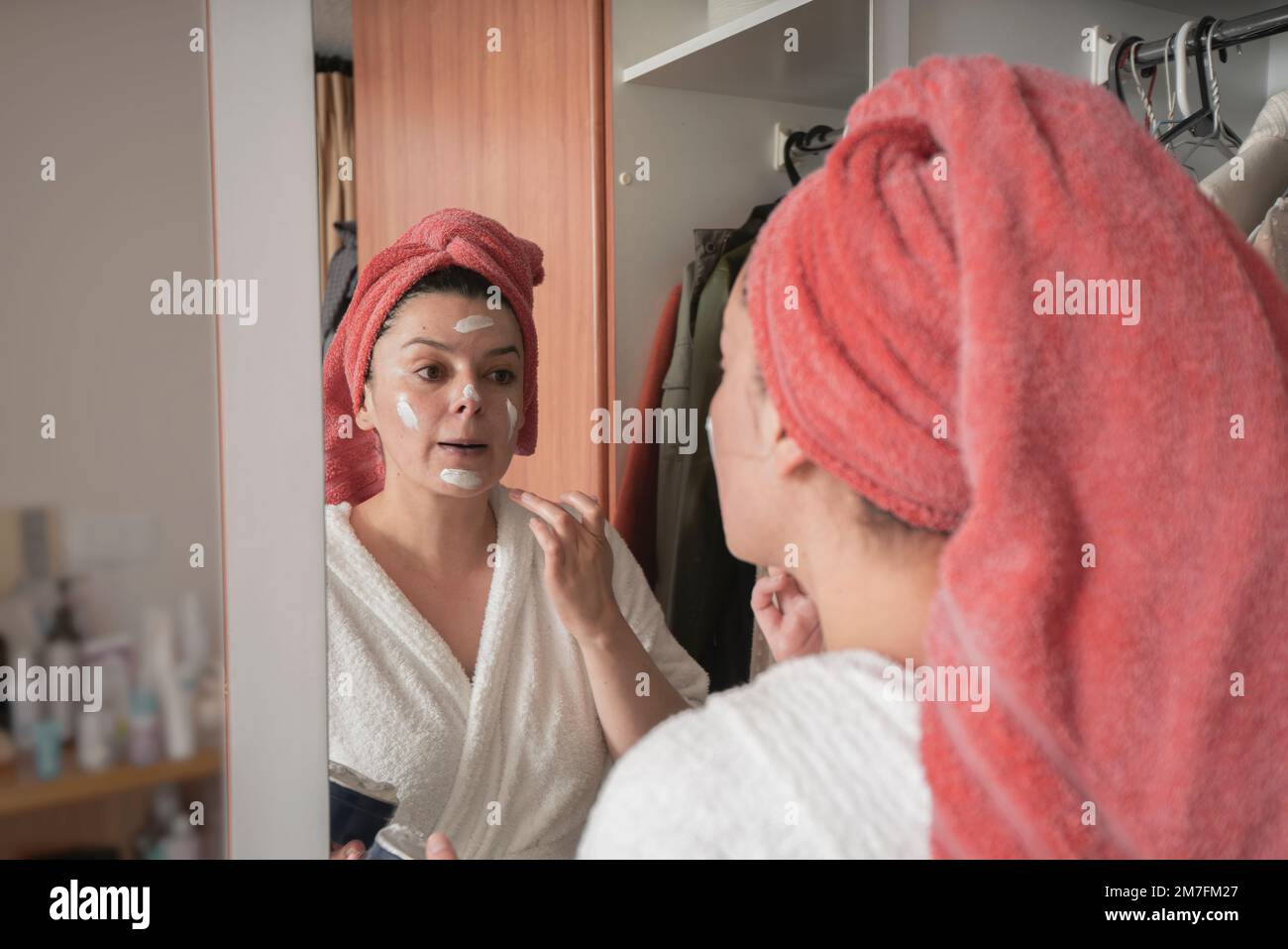Beautiful Hispanic woman in a white bathrobe and red towel in her hair applying makeup with her hand on her face in front of the mirror on the closet Stock Photo