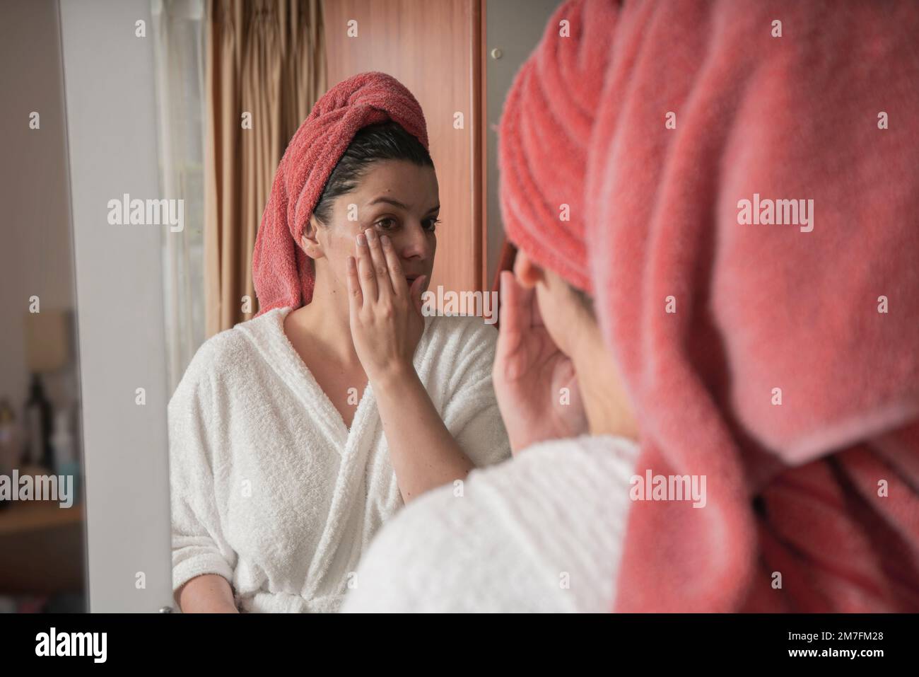 Beautiful Hispanic woman in a white bathrobe and red towel in her hair applying makeup with her hand on her face in front of the mirror on the closet Stock Photo