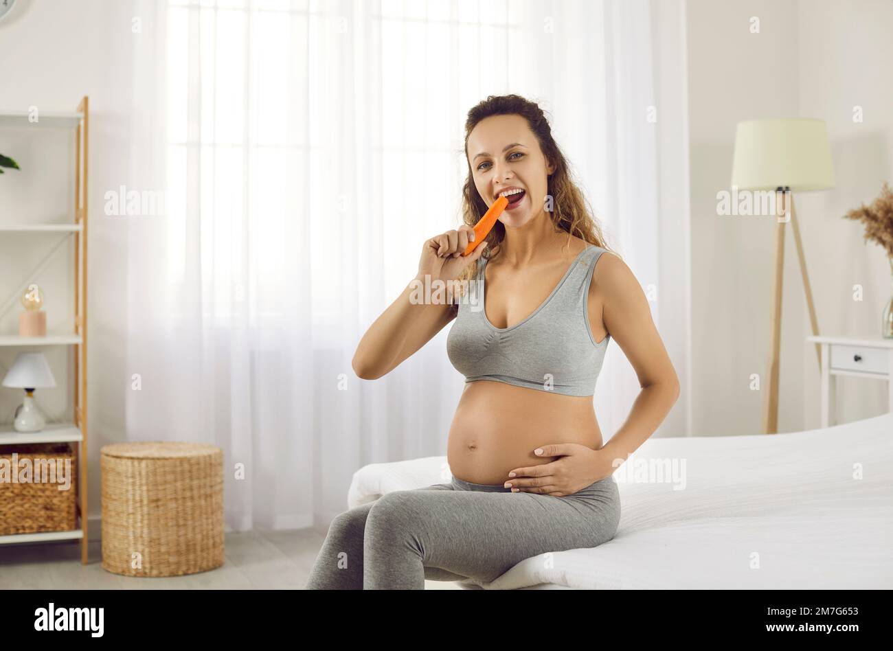 Happy young pregnant woman sitting in the bedroom and eating a fresh organic carrot Stock Photo