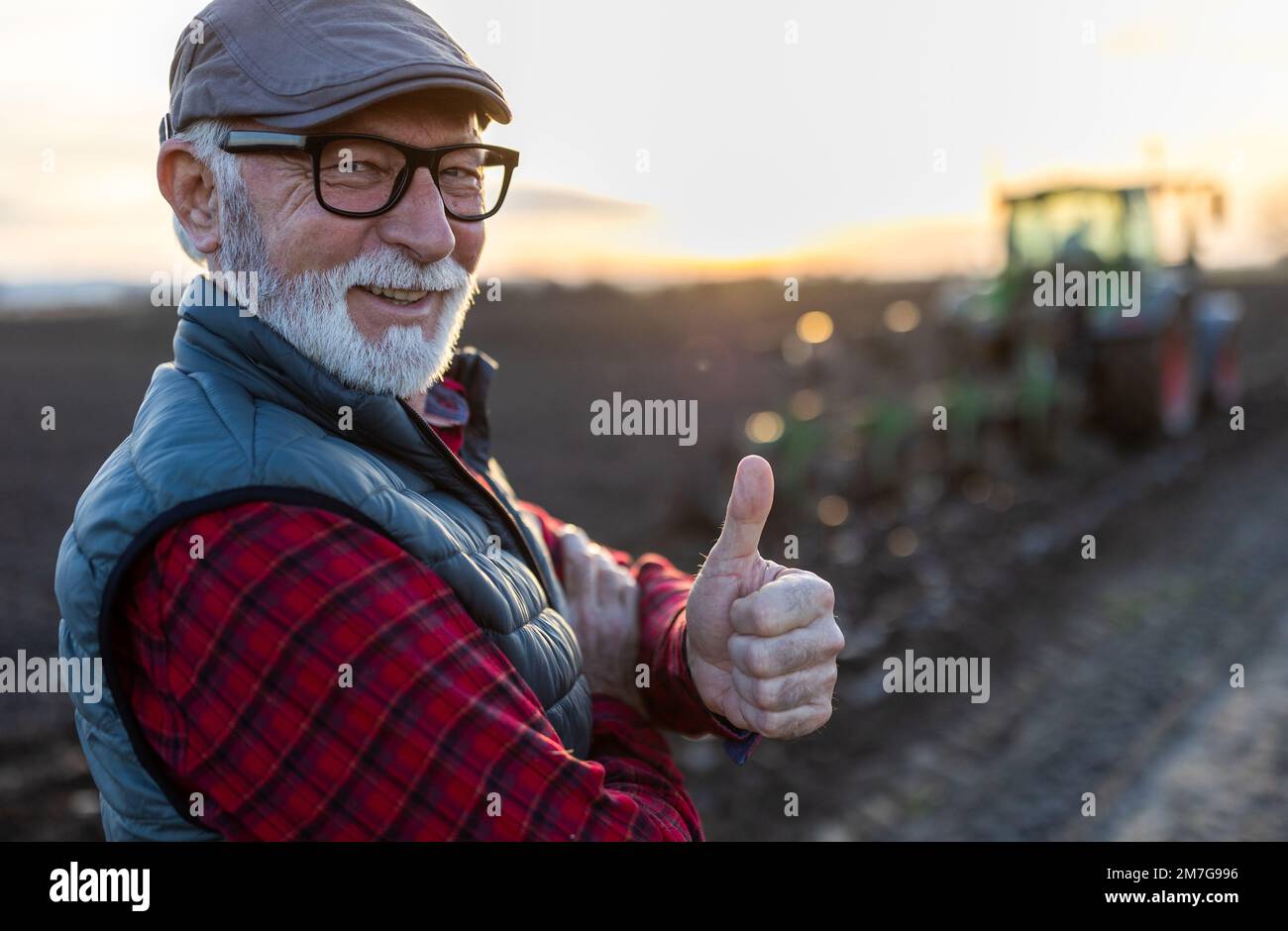 Portrait of mature farmer showing thumb up in front of tractor in field in autumn Stock Photo