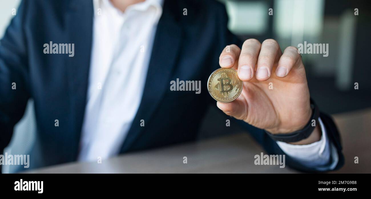 Closeup shot of businessman showing bitcoin, sitting at workplace in office, selective focus, panorama, copy space Stock Photo