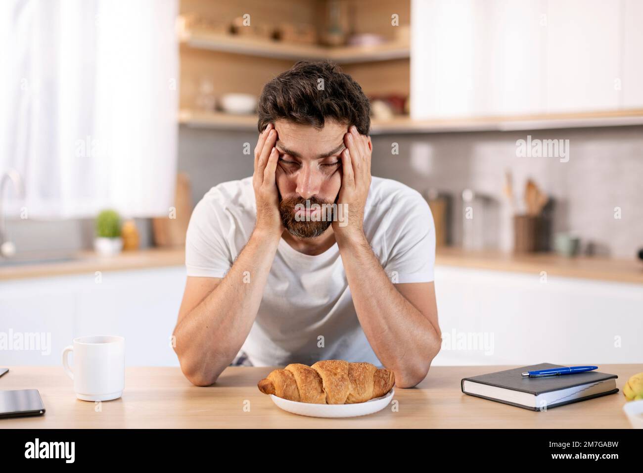 Despaired tired millennial caucasian man with beard holds head with hand, asleep at table with croissant Stock Photo