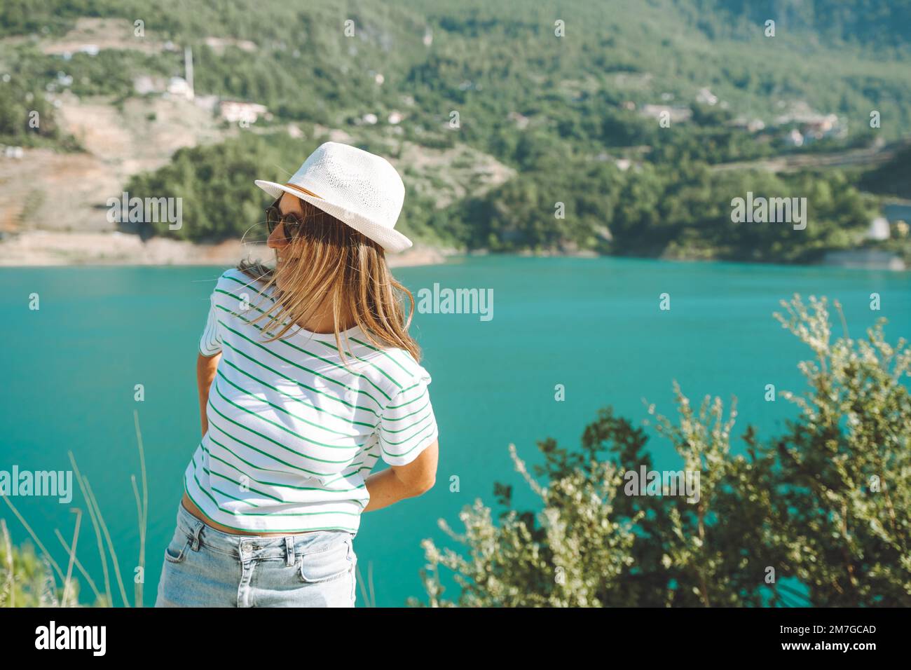Smiling woman in hat and sunglasses with wild hair standing near mountains lake on background. Positive young woman traveling on blue lake outdoors Stock Photo