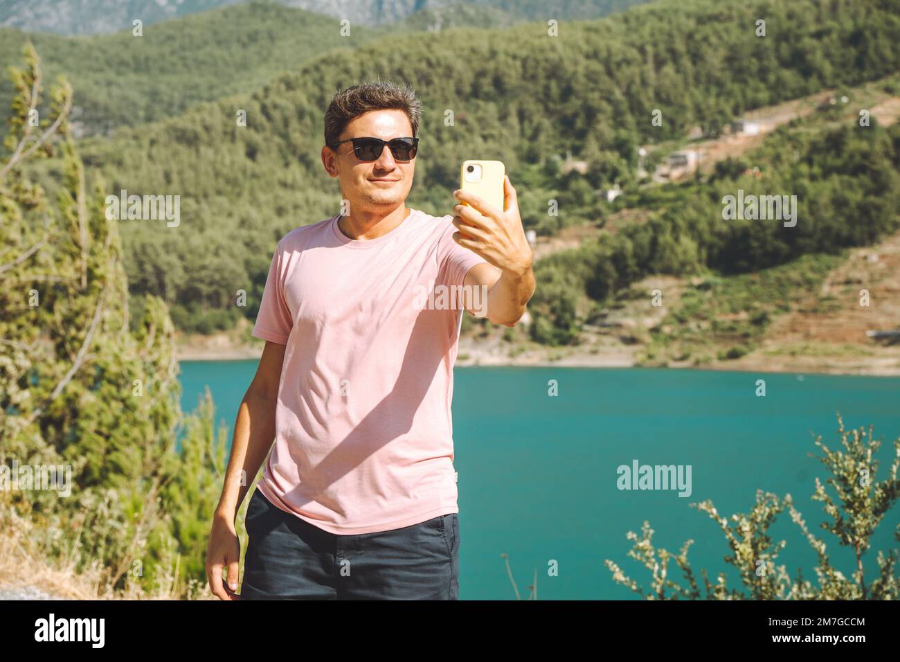 Man taking selfie on mobile phone of mountains lake background. Traveler male having video chat on cellphone on the blue lake outdoors travel Stock Photo