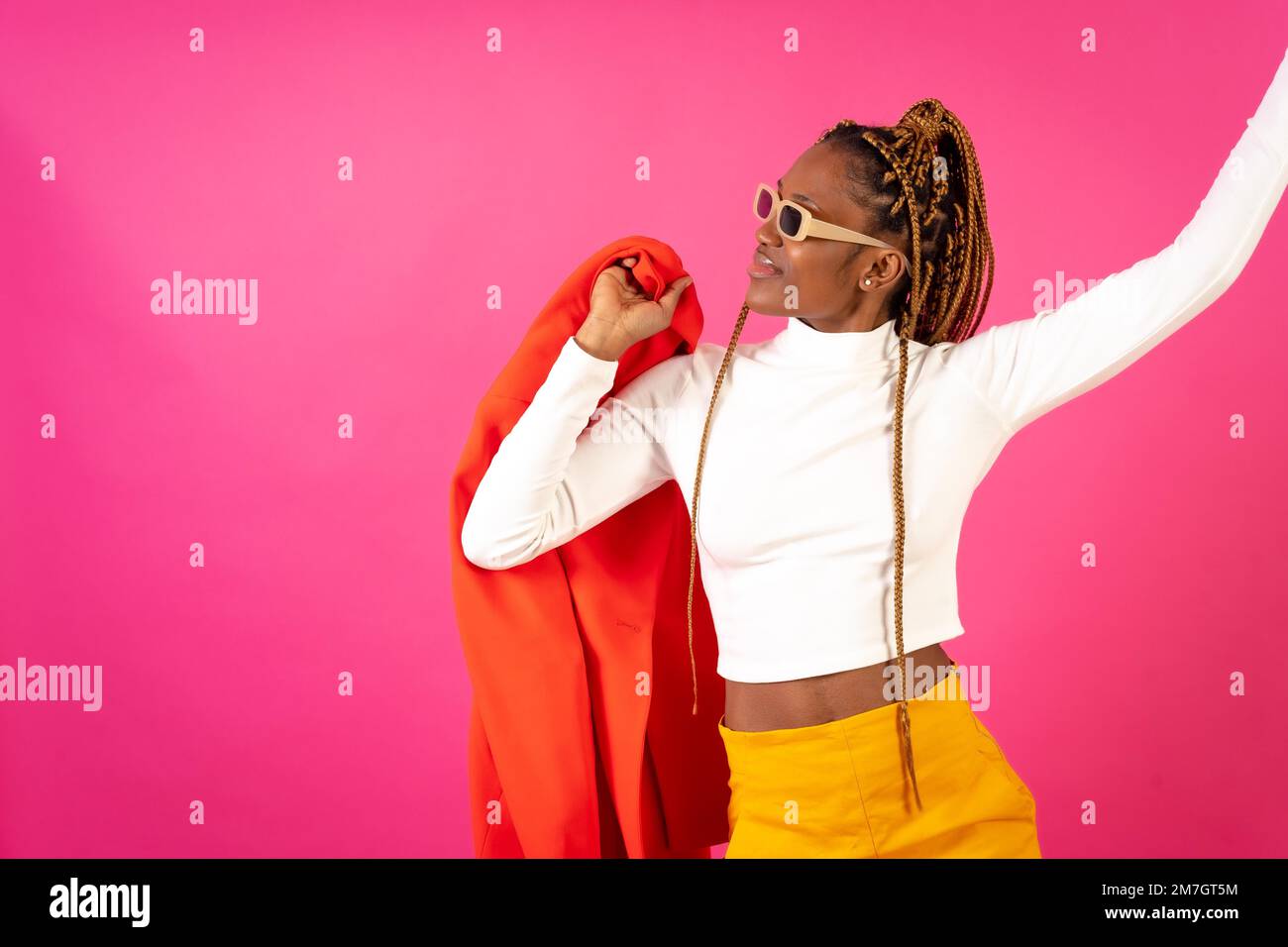 Black ethnic woman with braids on a pink background, wearing a red suit and white t-shirt dancing Stock Photo