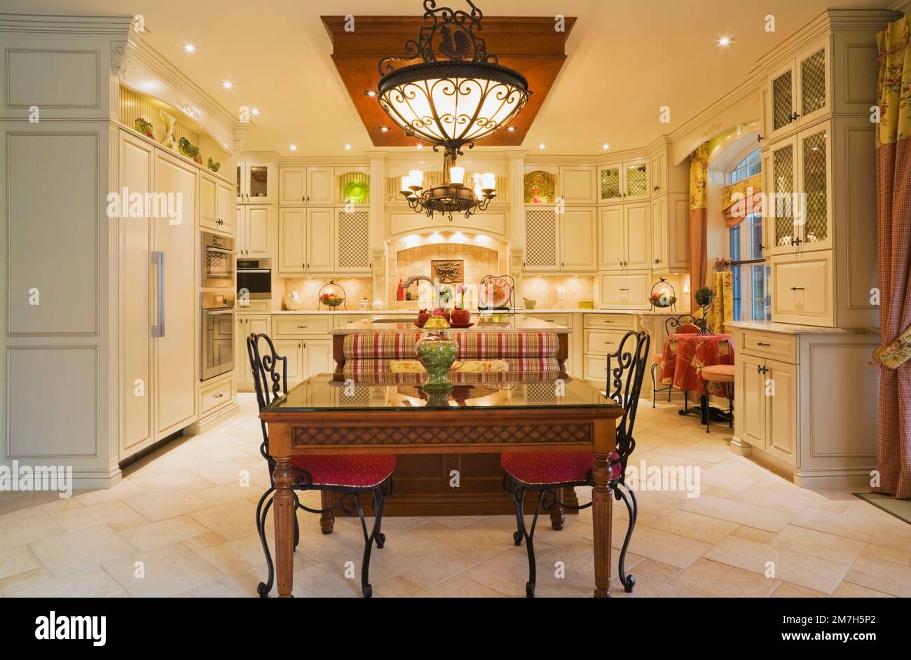 Illuminated chandeliers above breakfast table and red upholstered chairs in kitchen inside elegant cottage style home. Stock Photo