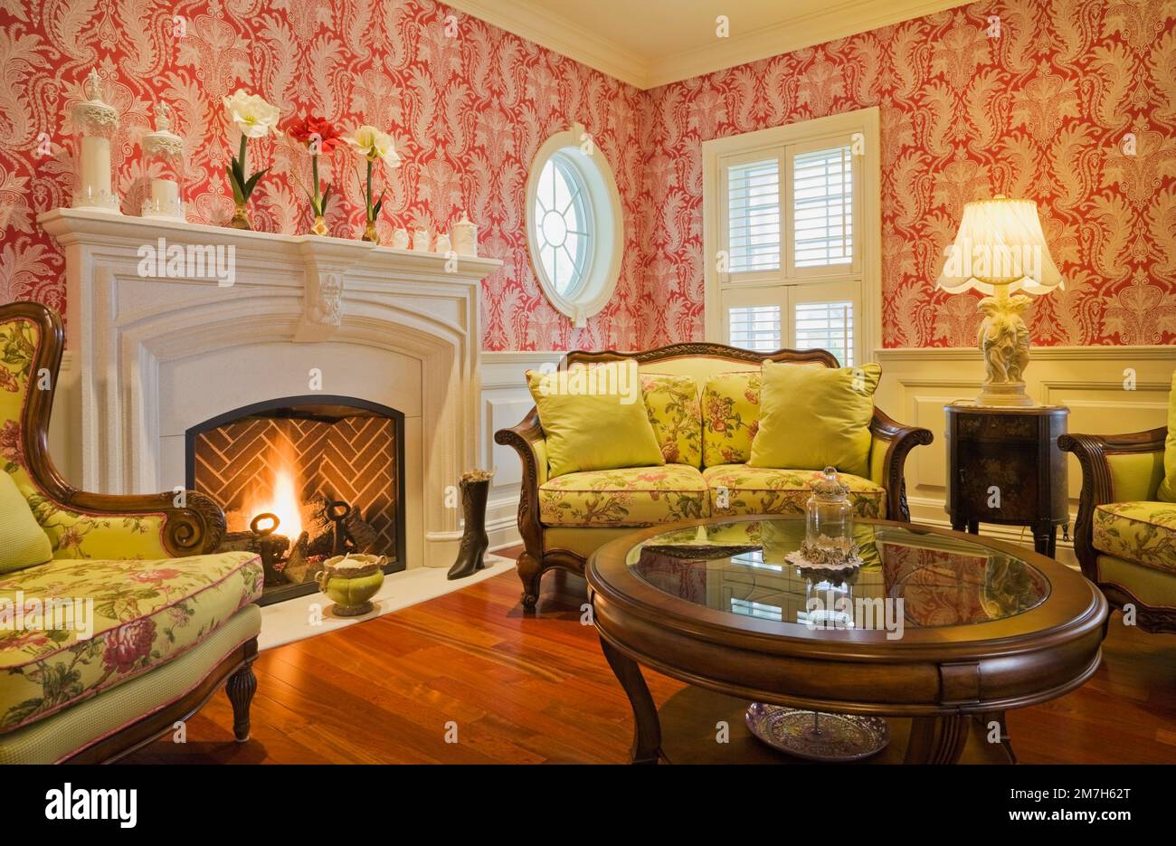 Lit propane gas fireplace and yellow flowery upholstered armchairs and sofa in parlour room inside elegant cottage style home. Stock Photo