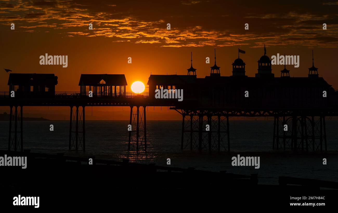 The golden orb of the rising sun appears between two buildings on the silhouetted Eastbourne Pier at dawn with clouds under-lit in the background Stock Photo