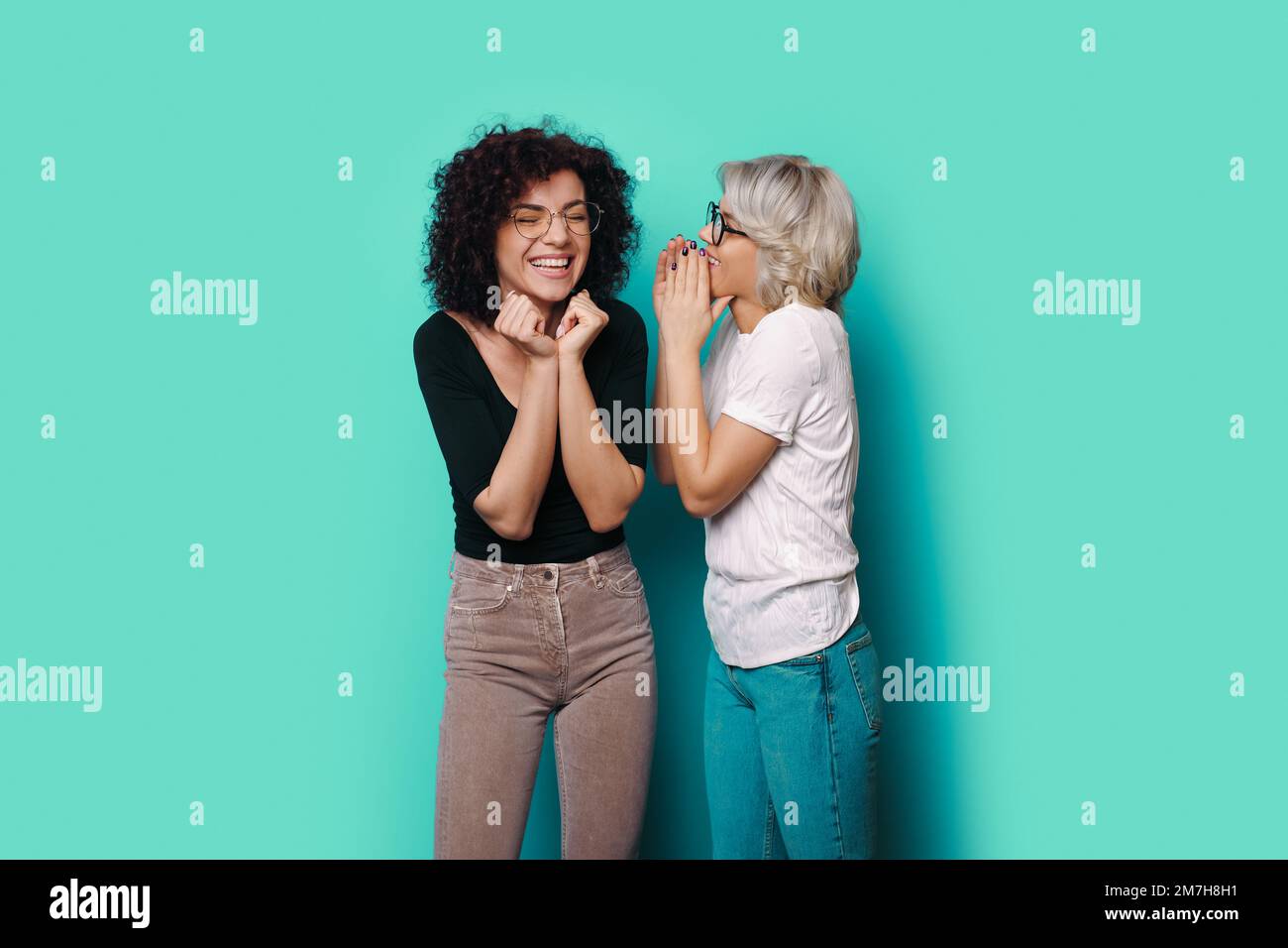 Excited woman listening to friend whispering to her ear on the blue background. Fashion model. Attractive beautiful girl. Stock Photo