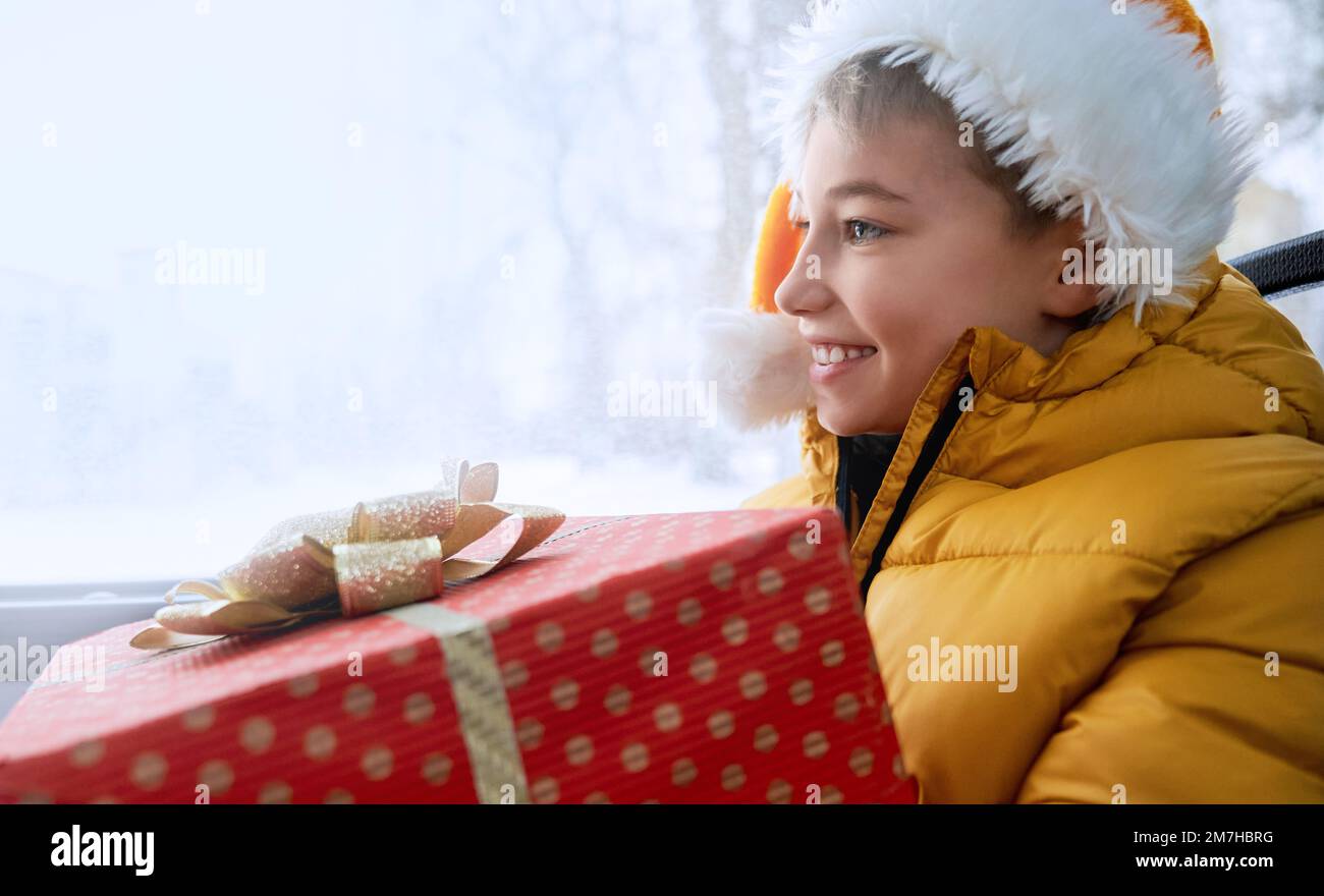 Side view of small kid wearing Christmas hat, traveling by public tarnsport, sitting on bus, smiling. Cute kid holding gift box, looking forward. Concept of winter holidays. Stock Photo