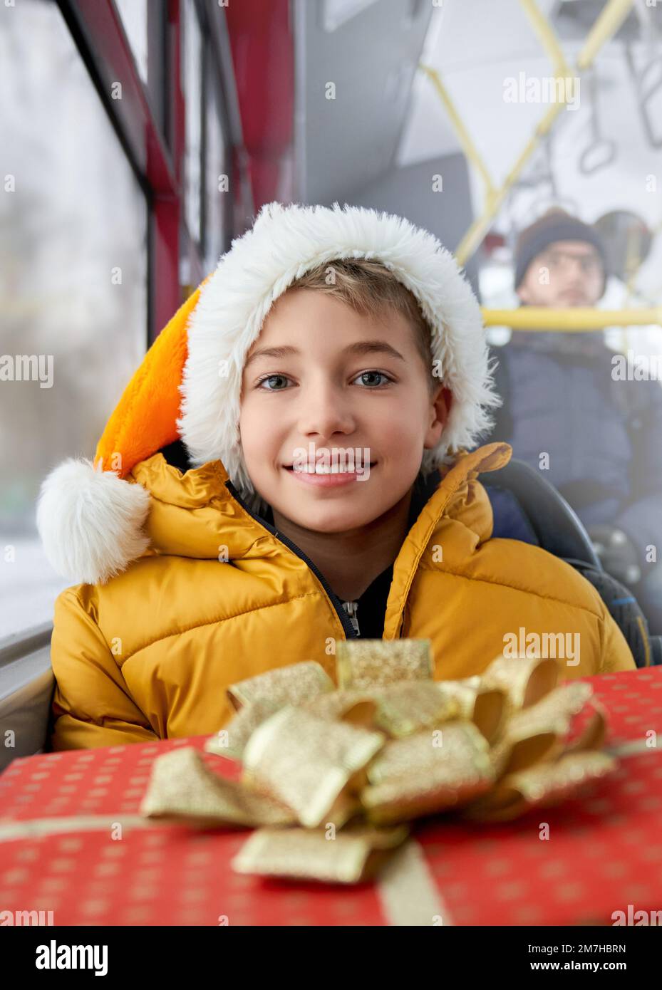 Front view of happy, cheerful kid traveling by public transport, bus during Christmas, winter holidays. Boy with Christmas hat sitting, looking at camera. Concept of winter vacations. Stock Photo