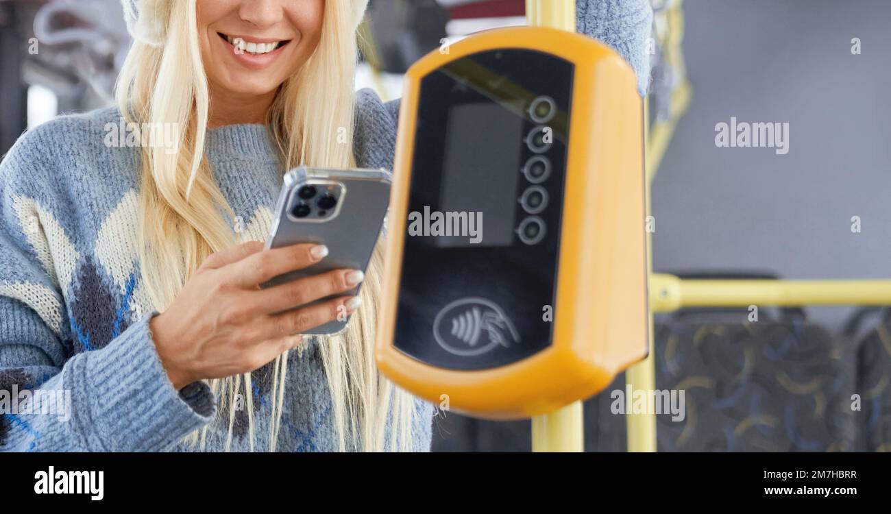 Crop of beautiful female wearing knitted sweater and neat manicure standing in bus performing contactless terminal payment. Getting to work by public transport coming through city. Concept of routine. Stock Photo