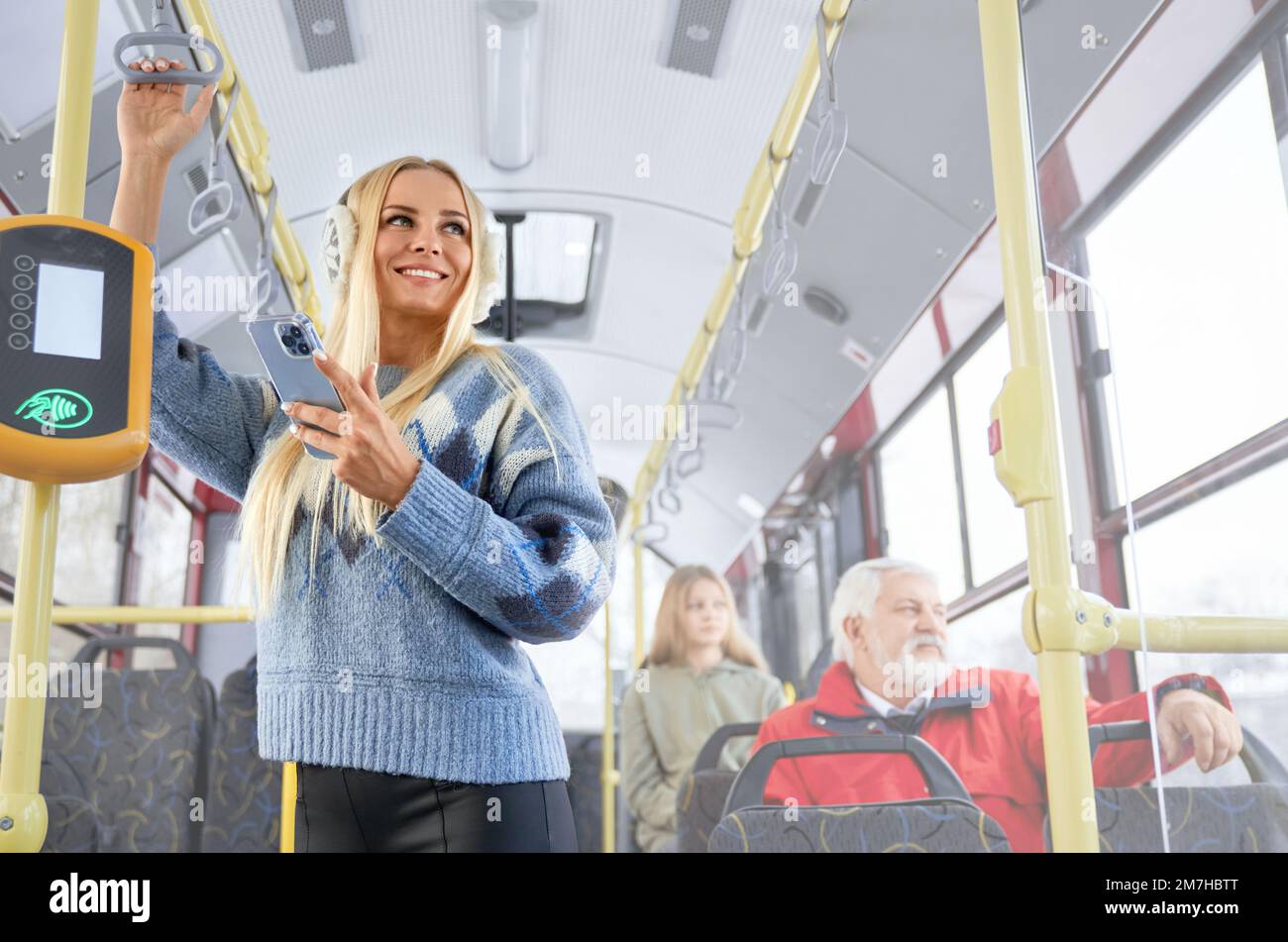 Portrait of pretty smiling woman standing in bus keeping holder and smartphone in her hands. Passengers of public transport looking through window outside. Concept of routine morning actions. Stock Photo