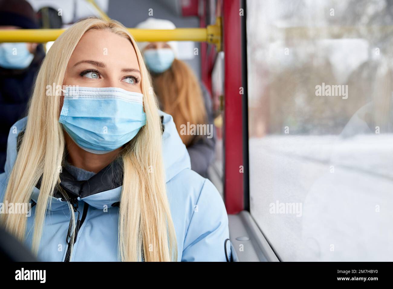Front view of blonde, attractive woman going home by public transport, wearing protecting mask. Passengers sitting on bus, protecting from coronavirus. Concept of everyday routine. Stock Photo