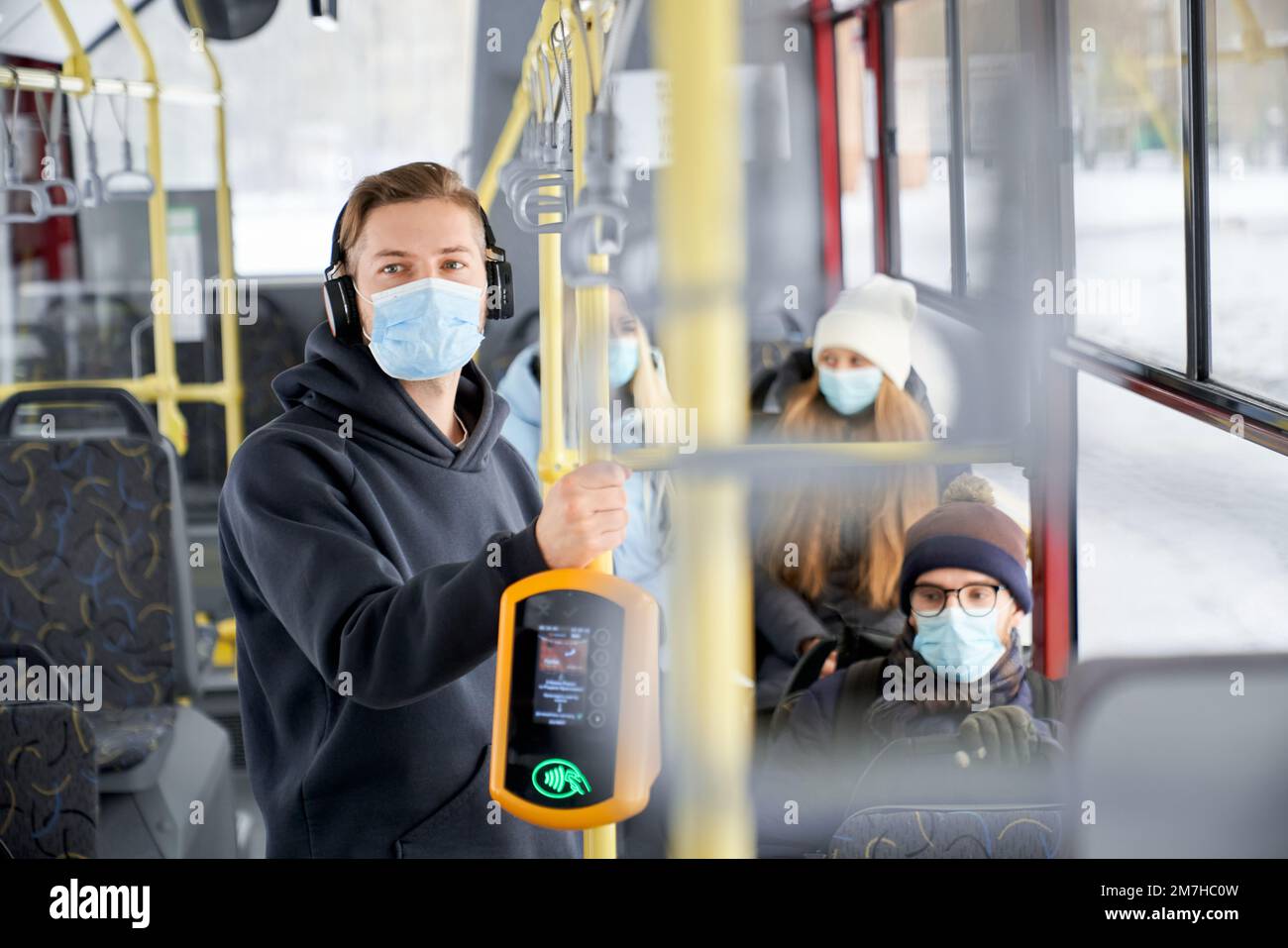 Side view of passengers standing, sitting on public transport, wearing medical masks, protecting. Young man holding handle, listening to music. Concept of quarantine measures. Stock Photo