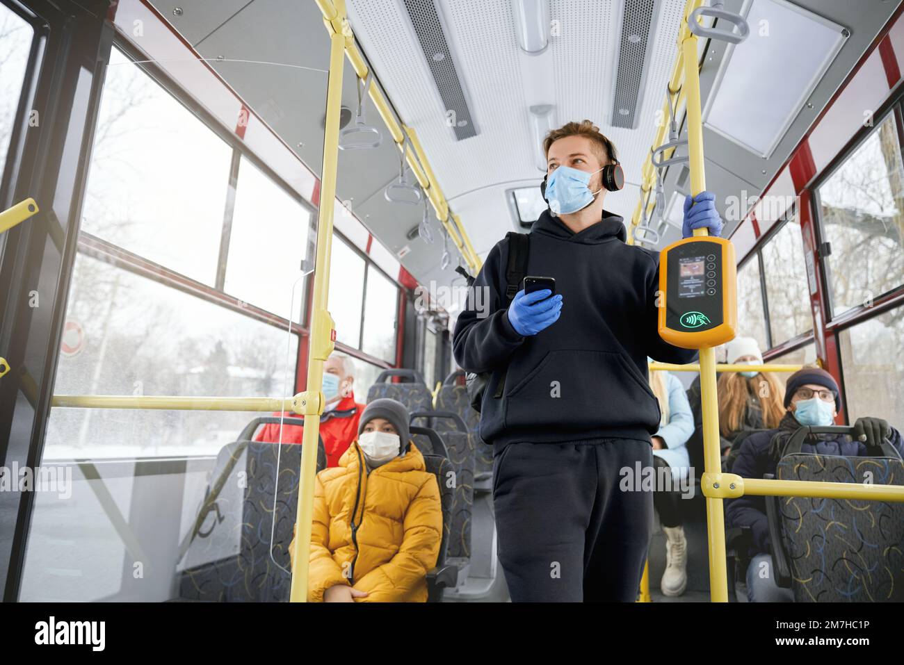 Front view of crowd traveling by public transport, wearing medical masks and gloves, protecting. Young brunette man with earphones paying by smartphone. Concept of quarantine. Stock Photo