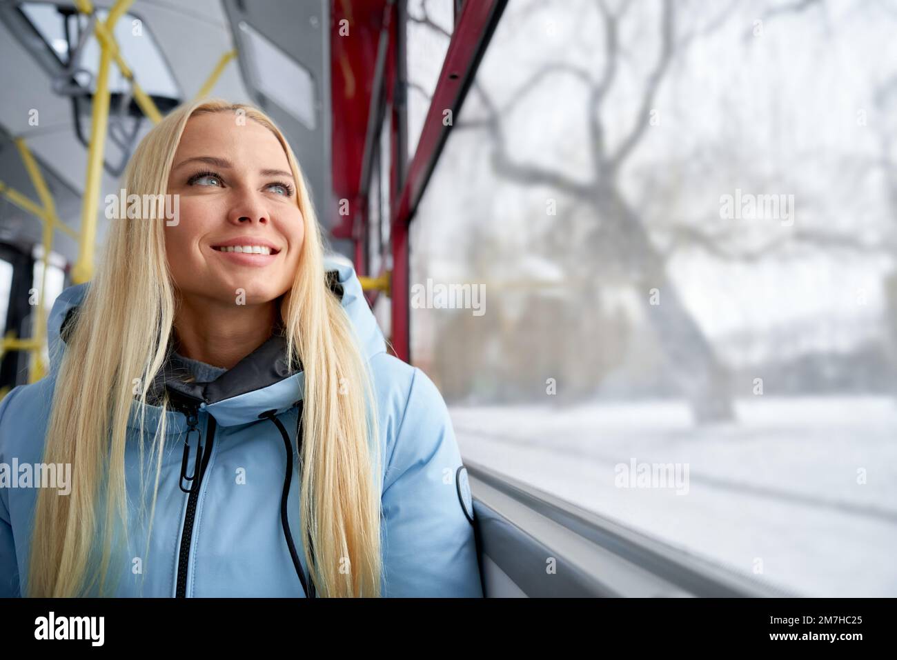 Front view of pretty, blonde woman with long hair sitting on bus, waiting, looking through window. Young female smiling, traveling by public transport. Concept of modern lifestyle. Stock Photo