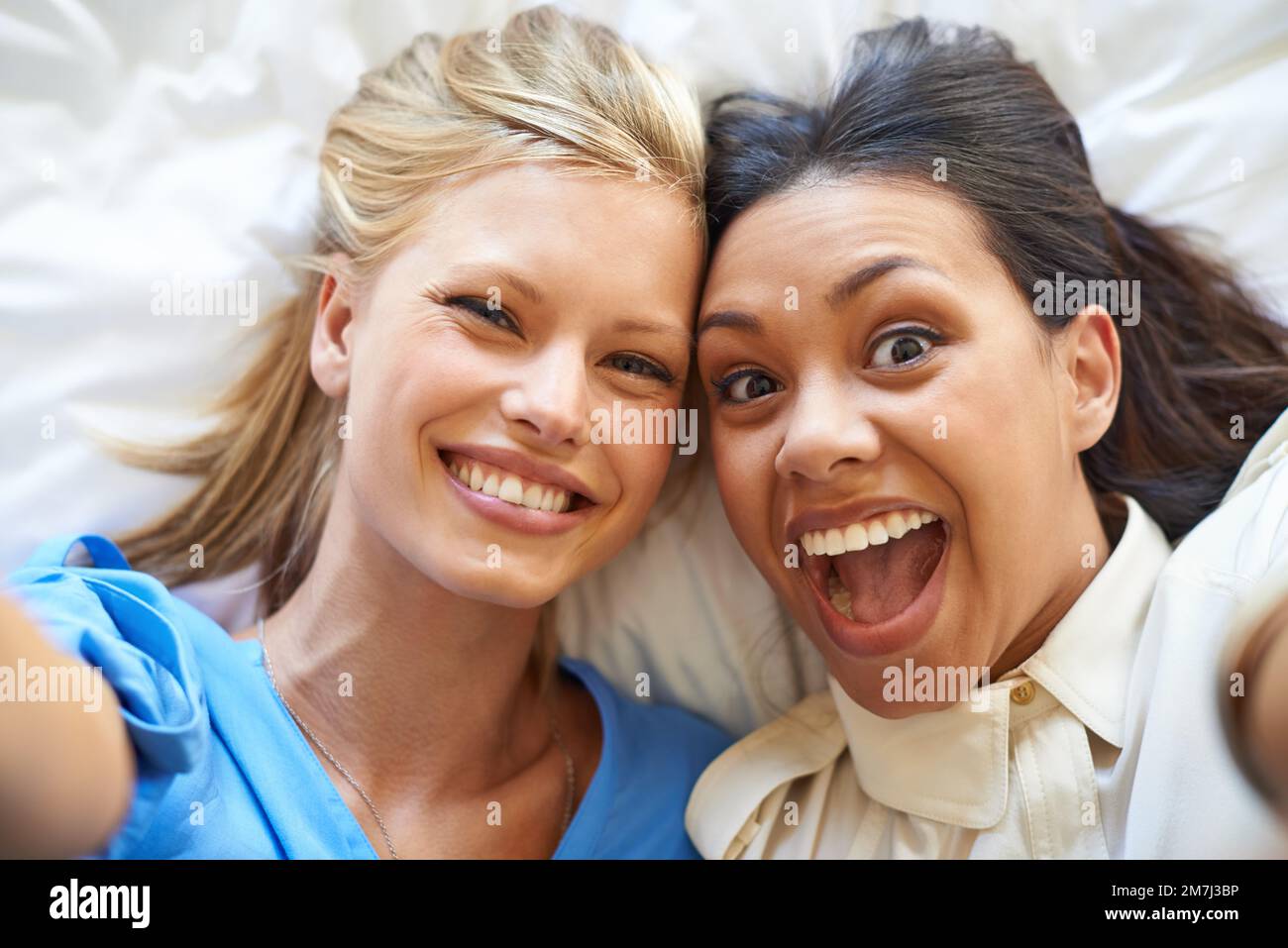Look happy. High angle portrait of two attractive young women taking selfies while lying on a bed. Stock Photo