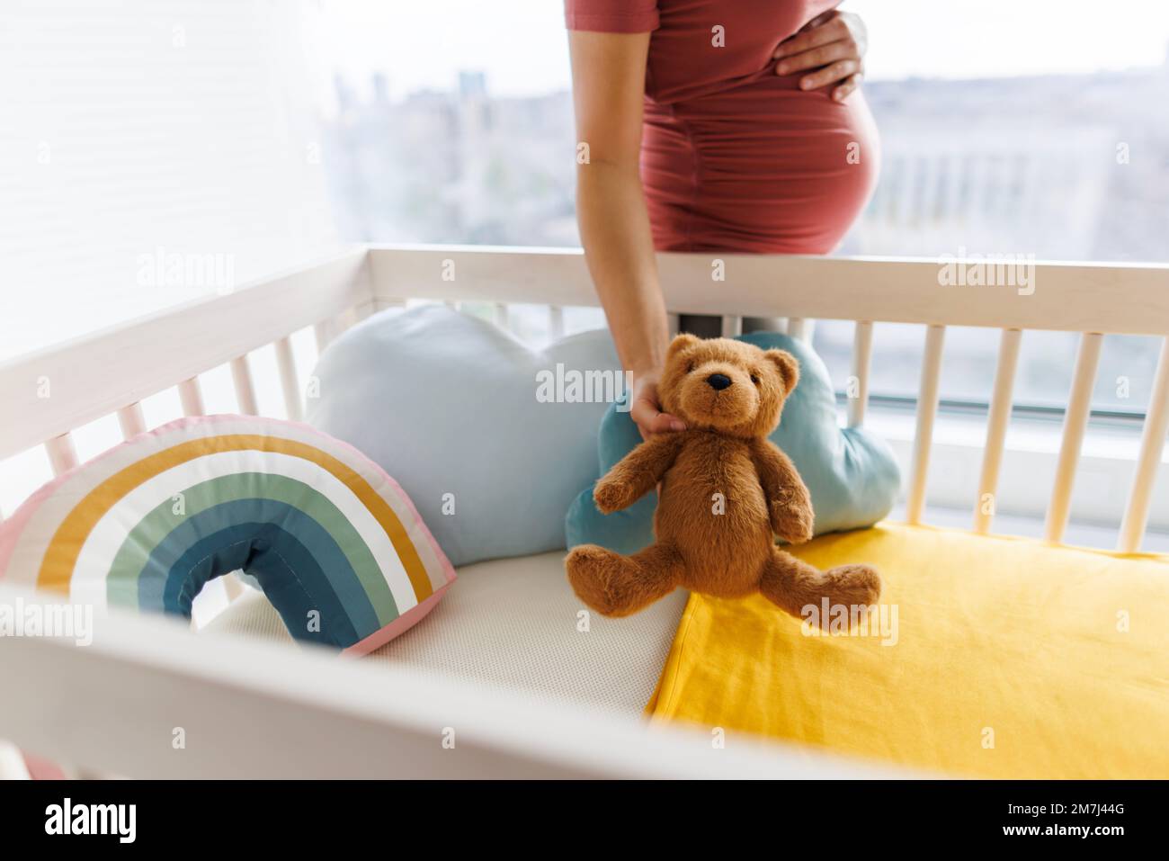 Pregnancy. Pregnant woman preparing nursery holding belly baby bump by crib holding teddy bear. regnancy concept and home nusery planning Stock Photo