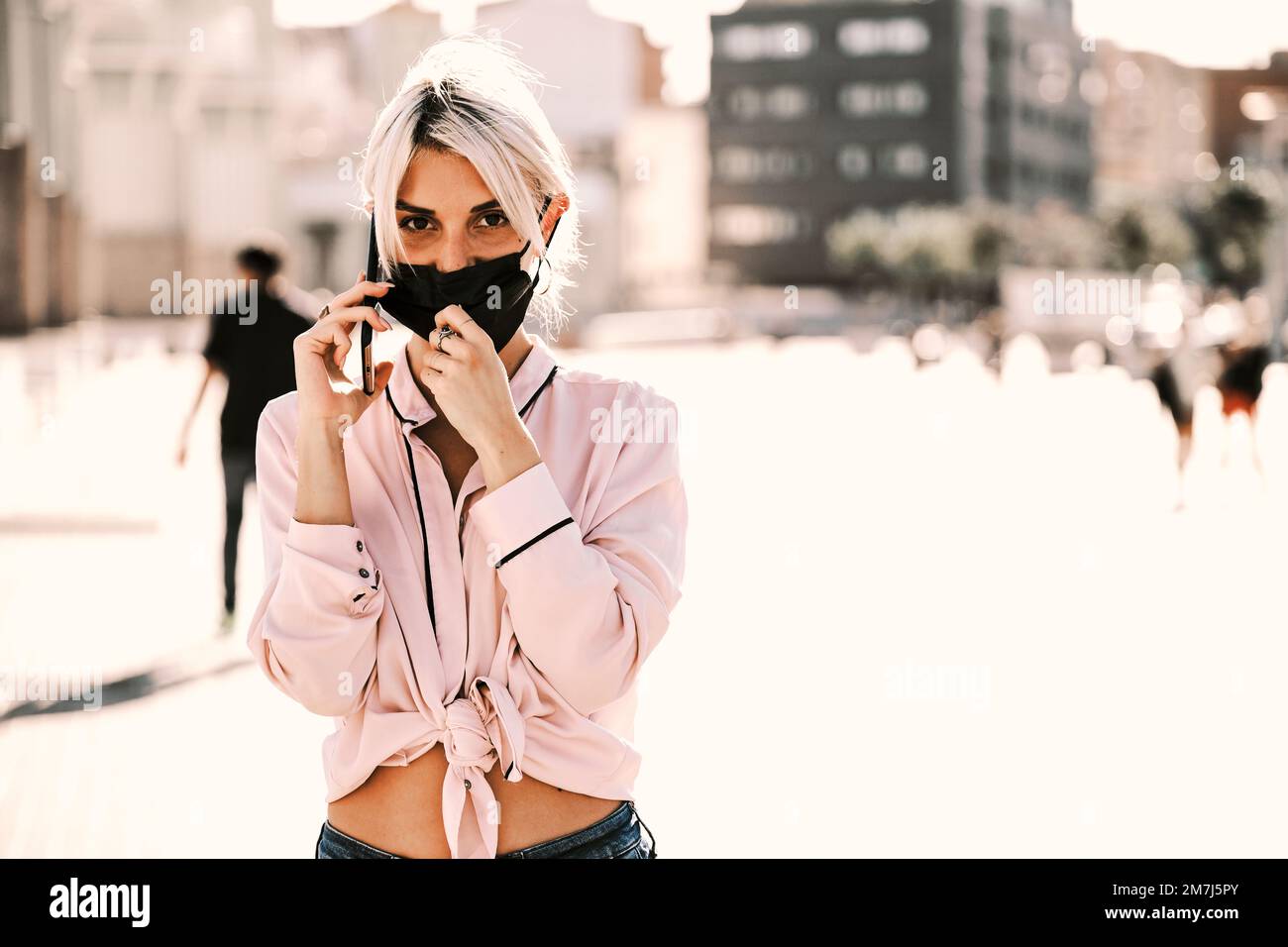 Woman with face mask looking at camera while talking on the phone outdoors on the street. Stock Photo