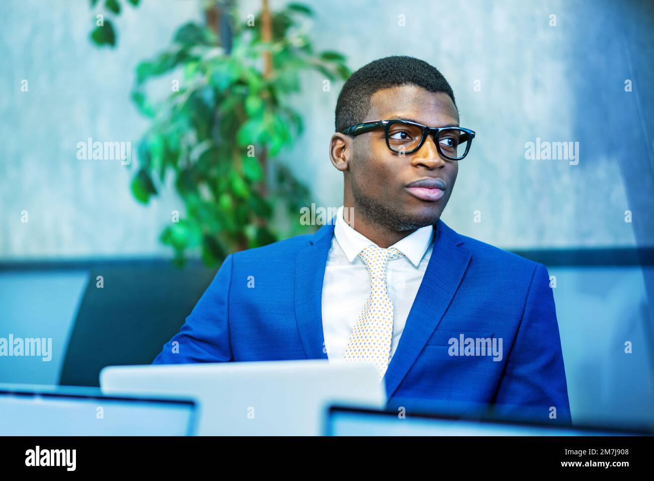 African American male entrepreneur in blue suit and stylish glasses looking away while using laptop during work in modern office Stock Photo