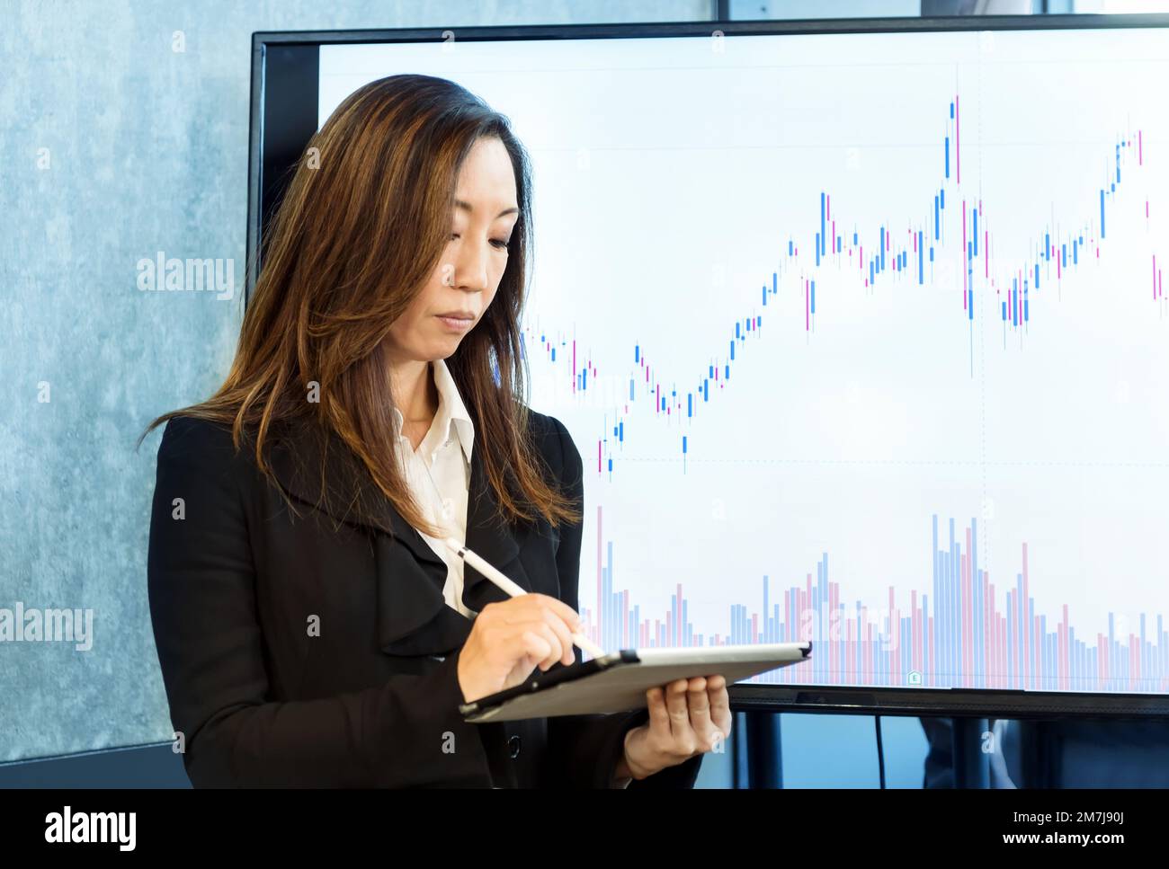 Concentrated ethnic female executive in formal suit standing near big monitor with financial chart while using tablet in office Stock Photo