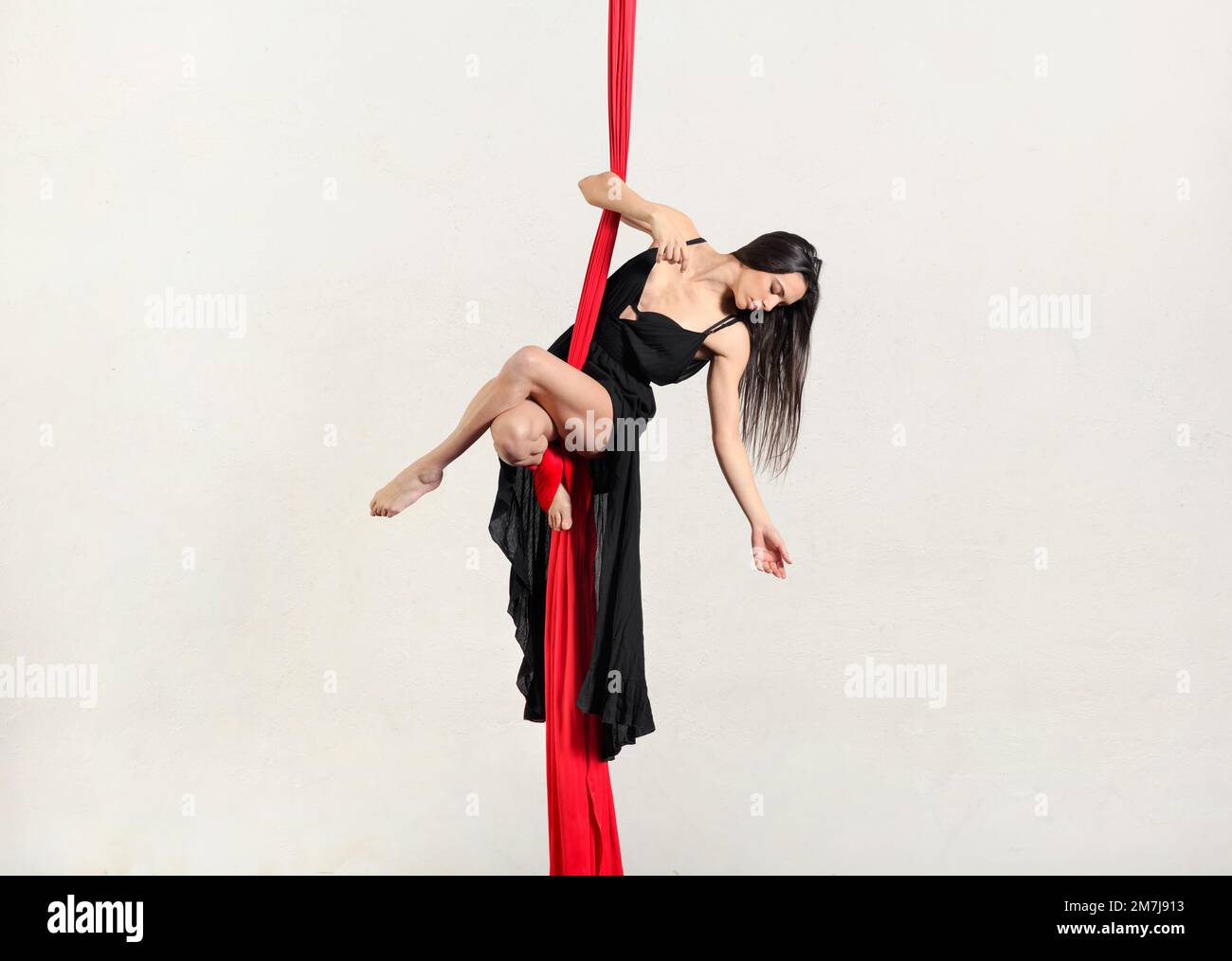 Full body of graceful female with long hair in black dress hanging on aerial red silks while doing yoga on white background Stock Photo