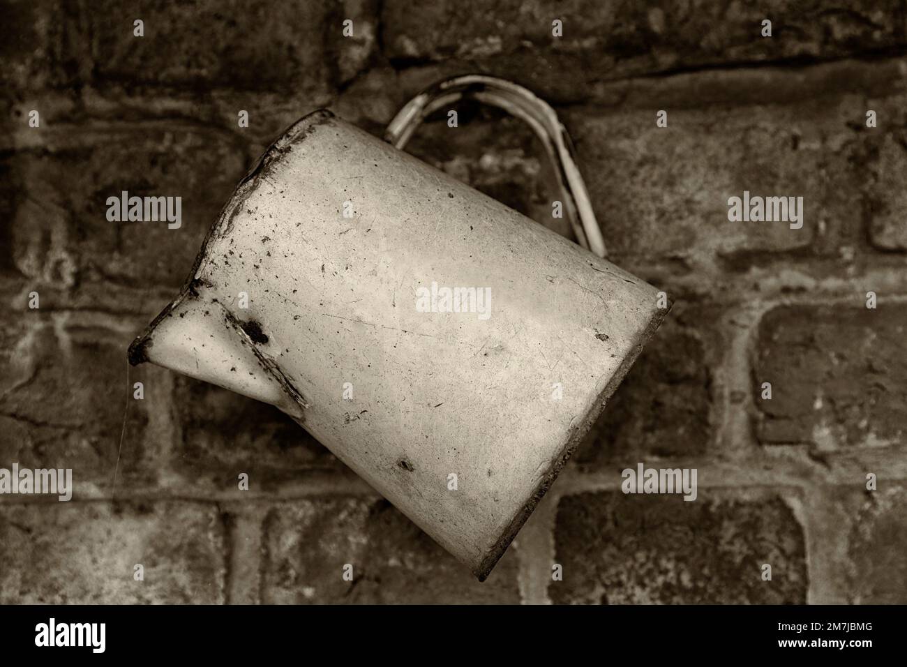 Black and white sepia toned image of an old rusty enamel water jug hanging on a wall Stock Photo