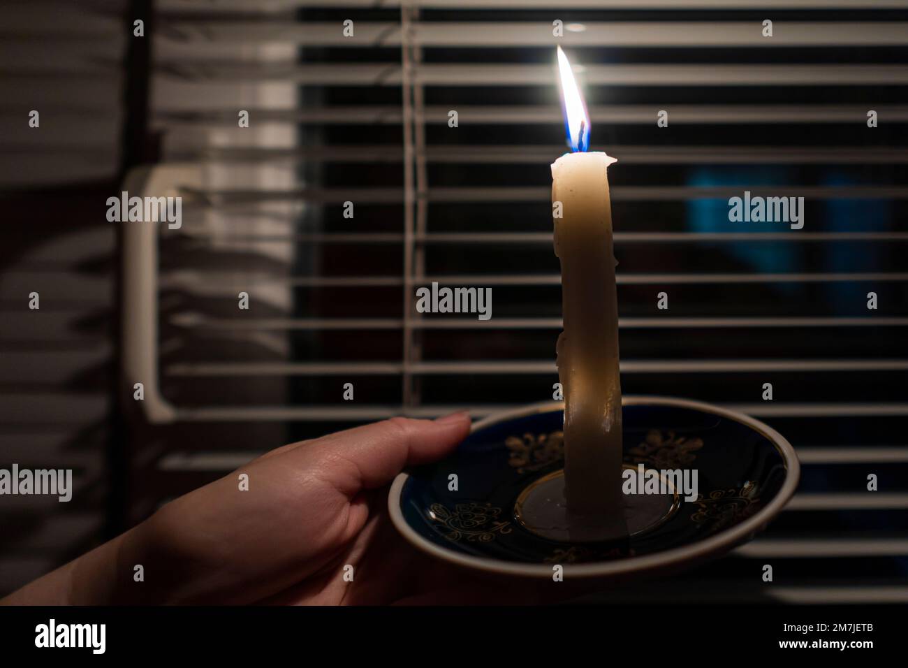 Burning candle on a saucer in the hands against the background of a window with blinds (close-up). Power outage concept. Blackout. Energy crisis Stock Photo