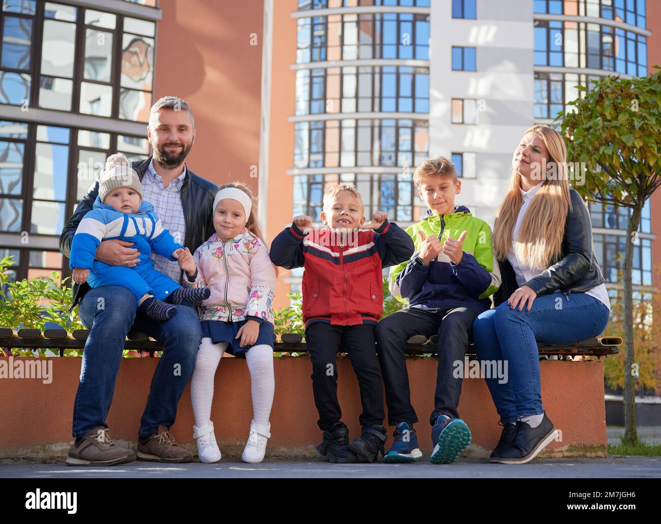 Happy family - father, mother and children having fun together on playground. Parents and kids sitting on bench, looking to camera. Modern residential buildings on background. Stock Photo