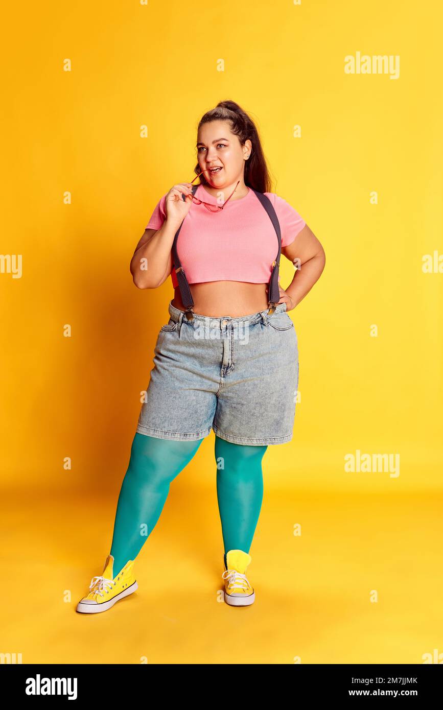 Portrait of young overweight woman in casual summer bright clothes posing over vivid yellow studio background Stock Photo