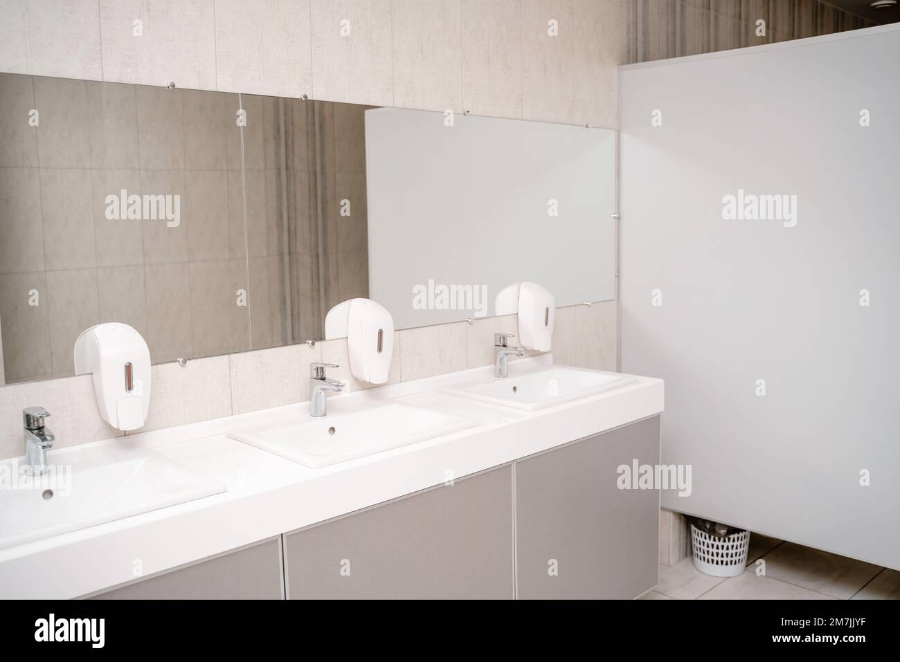 The interior of a public toilet. Row of wash basins with metal faucets on a marble slab, liquid soap dispensers, a long mirror on a gray wall Stock Photo