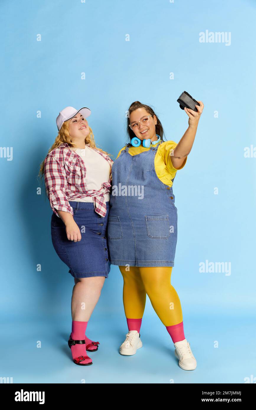 Portrait of two overweight women, friends taking photo, selfie with camera, posing over blue studio background Stock Photo