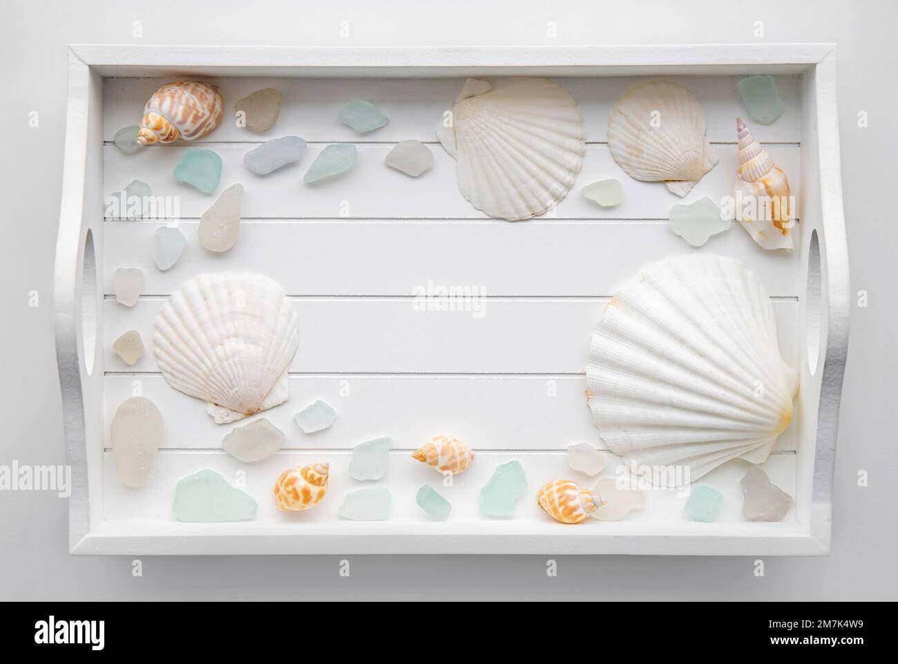 Flat lay view of white wood tray with seashells and sea glass, copy space in the center. Travel destination or beauty products background. Stock Photo