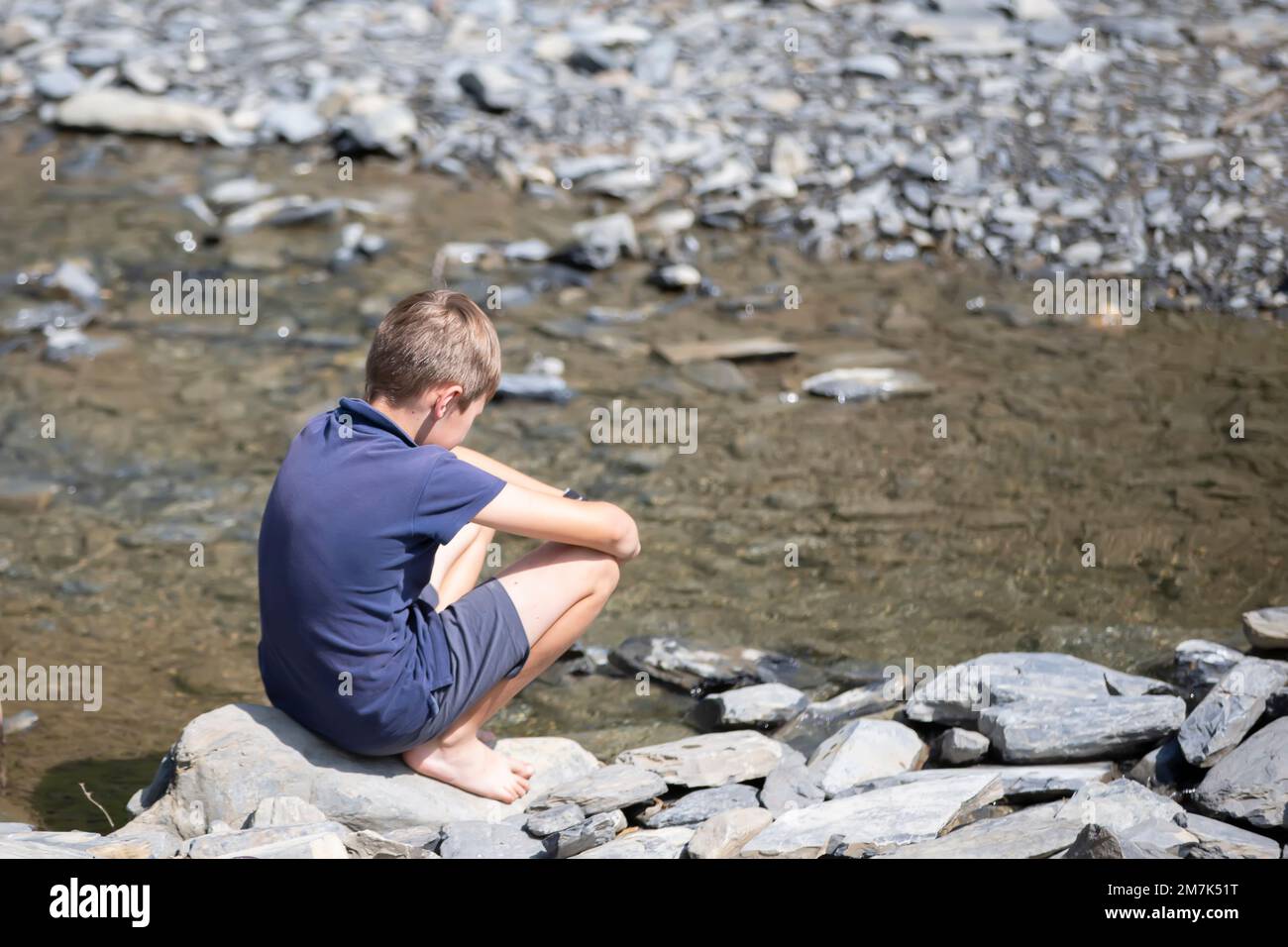 Little boy sitting on rocks with his back looking at the mountain lake ...