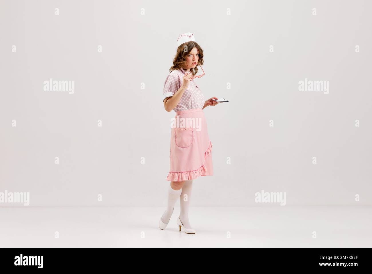Beautiful young girl in image of retro cater waiter wearing 70s, 80s fashion style uniform isolated over light studio background. Charm, ad, sales Stock Photo