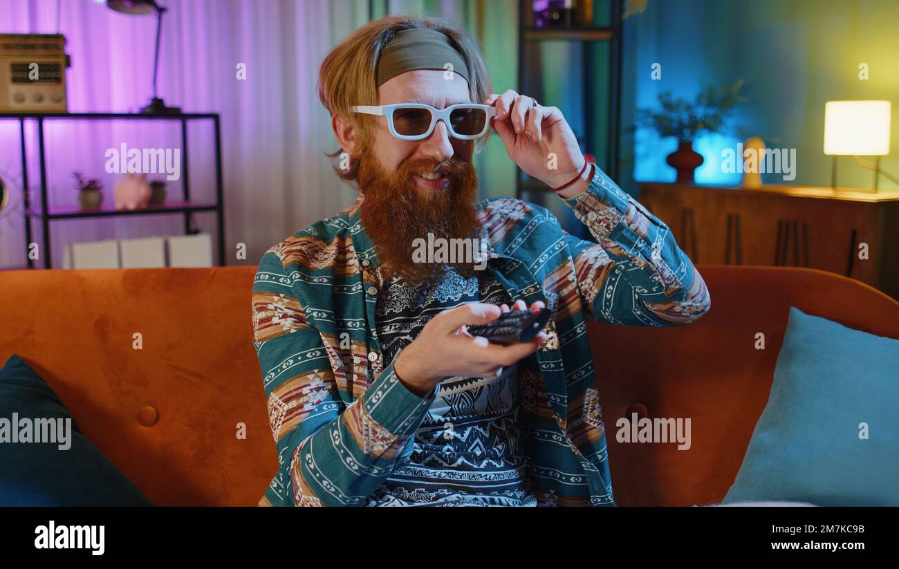 Excited bearded hippie young man sits on sofa use TV remote control watching serial, sport game,