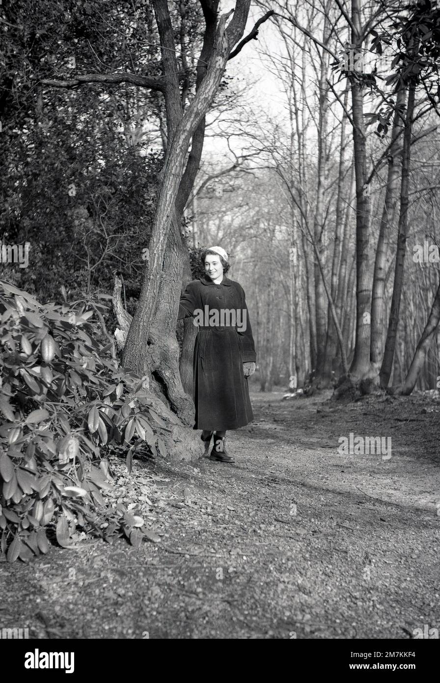 1950s, historical, a young lady wearing a long corduroy coat, small hat, gloves and ankle boots standing by a tree on a path in woods near Farningham, Kent, England, UK. Stock Photo
