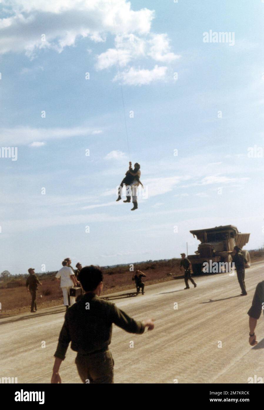 DF-SD-03-17789. [Complete] Scene Caption: AIRMAN First Class (A1C) William Hart Pitsenbarger, Pararescue Crew Member, Detachment 6, 38th Aerospace Rescue and Recovery Squadron (ARRS) lowered onto a roadway after an extraction of an Army of the Republic of Vietnam (ARVN) soldier from a burning minefield at Bien-Hoa Air Base for immediate medical attention. The soldier lost a foot when he stepped on a landmine. Nobody could figure out how to extract the wounded soldier without tripping the mines. A1C Pitsenbarger said, 'No problem, just lower me down on the penetrator, Ill straddle the guy, pick Stock Photo