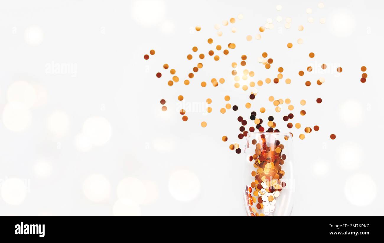 Top view on transparent wine glass with exploded firework of golden spangles. Festive copy space with crockery on white background with light bulbs. Stock Photo