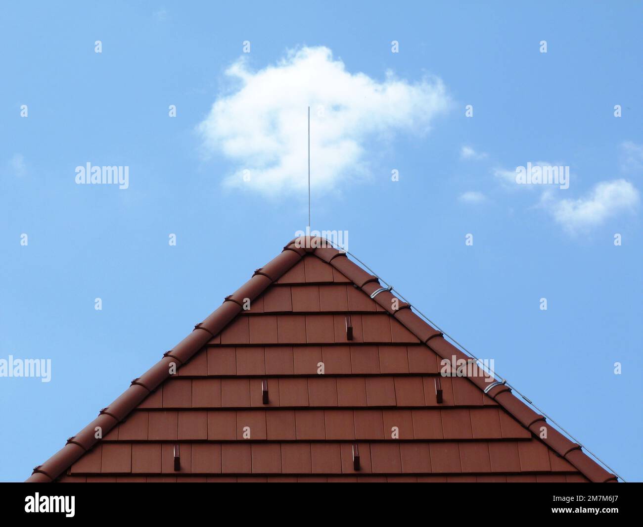 brown clay tile sloped pitched roof. metal snow breakers. ridge tiles and hip. steel lightning rod. roof construction materials. blue sky with clouds Stock Photo